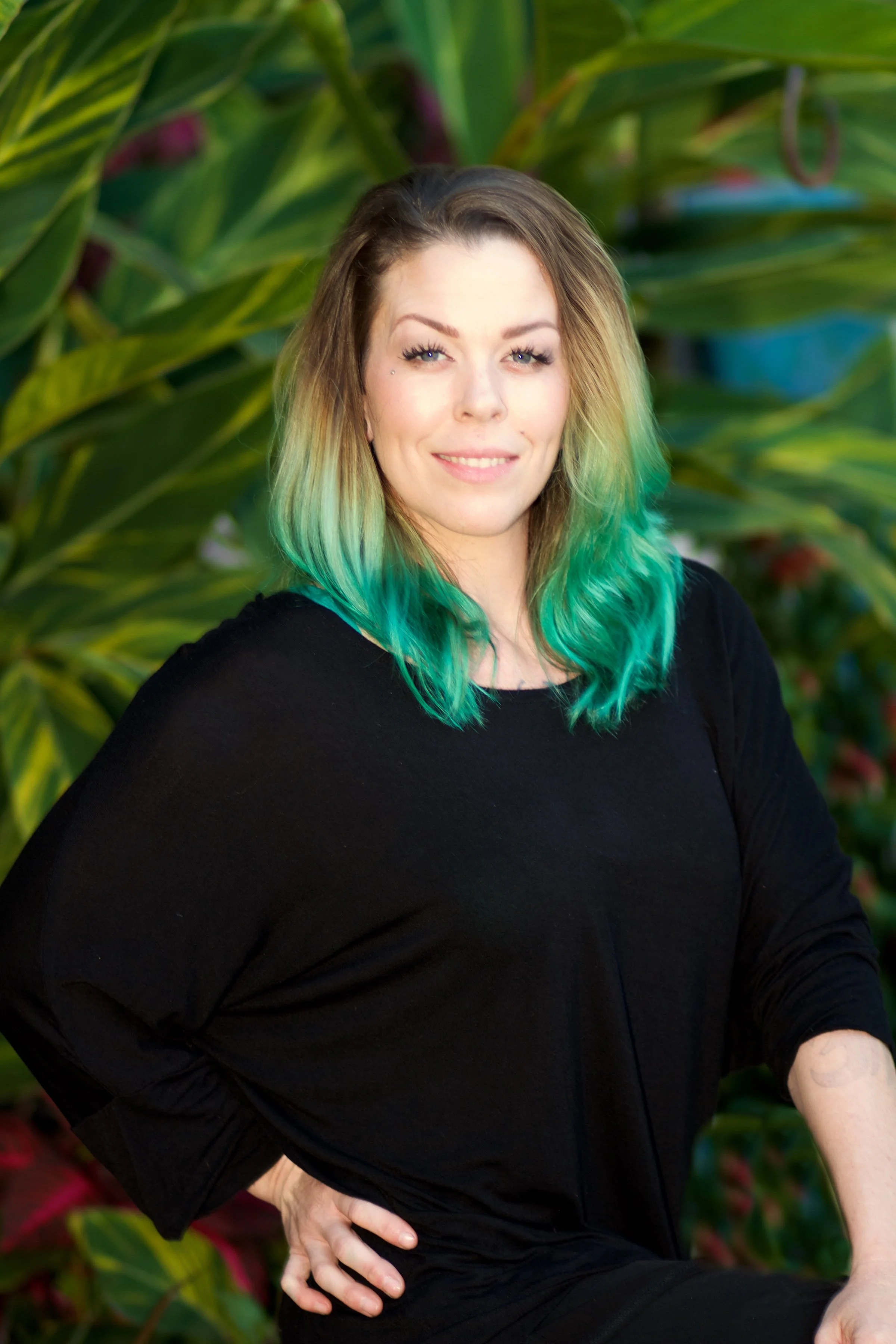 A white woman with shoulder-length blonde hair with green tips, wearing a black shirt, standing outdoors with green leafy plants in the background.