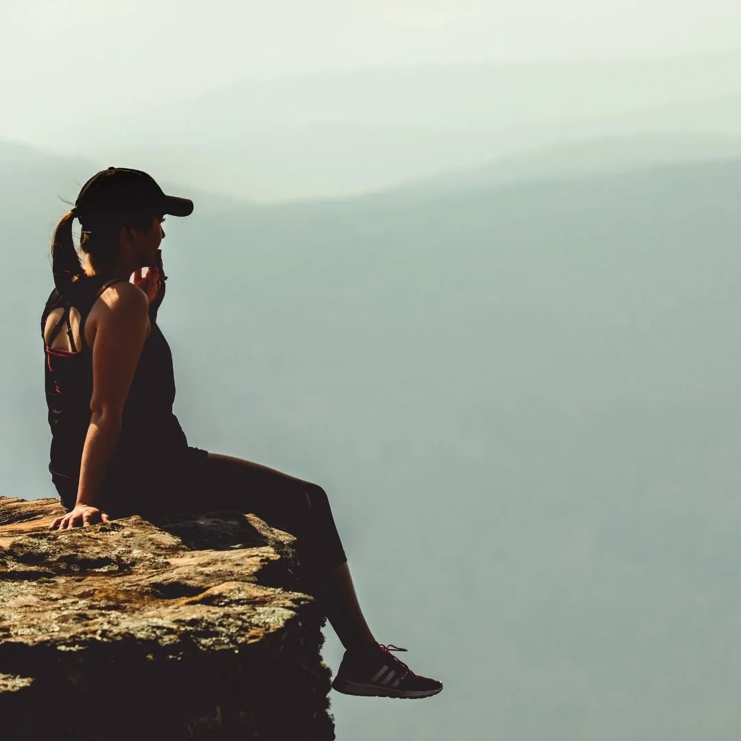 Silhouette of a woman sitting on a rocky ledge at the edge of a cliff, looking out over a foggy or cloudy landscape, wearing a cap and athletic clothing.
