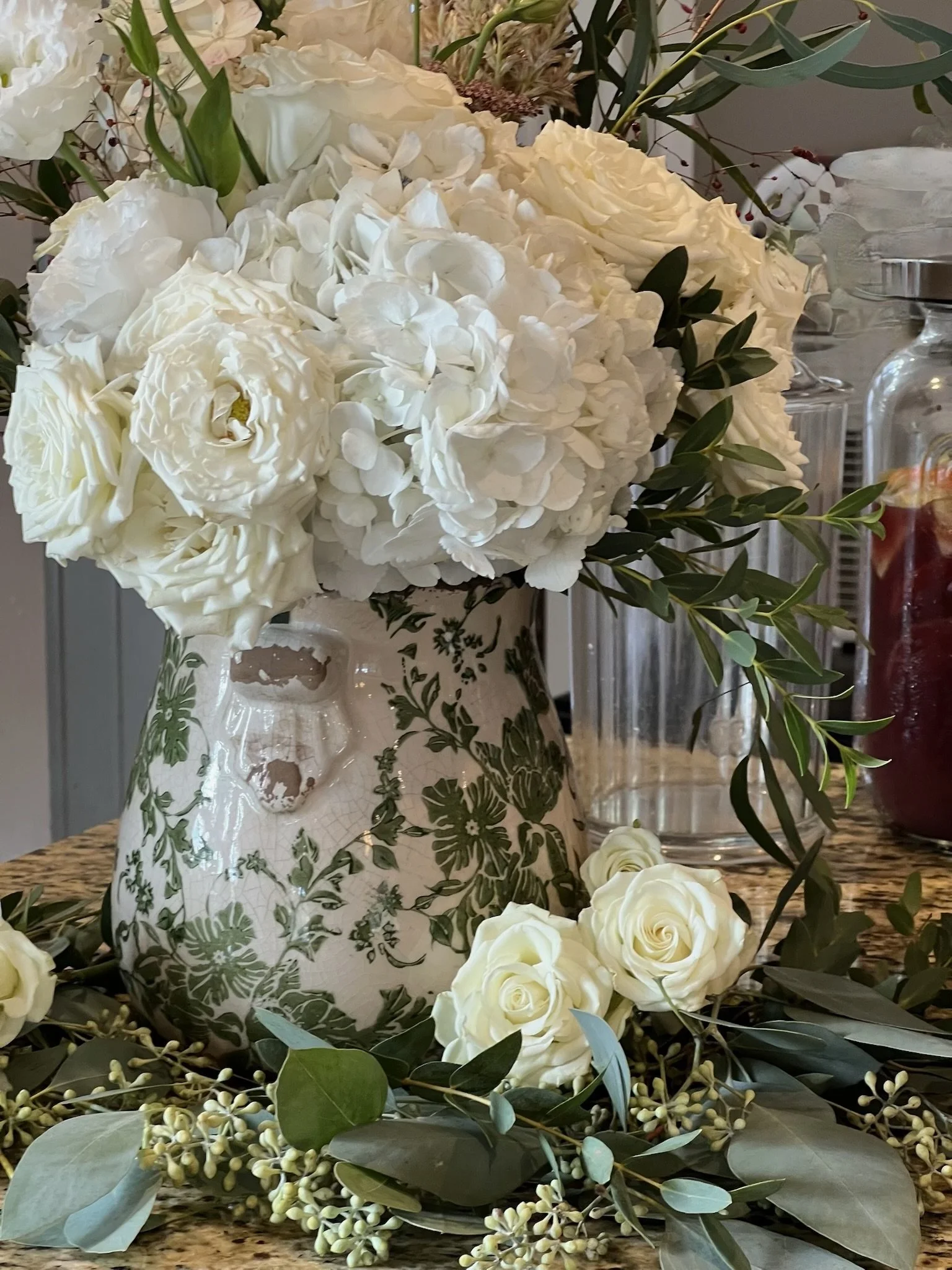 A decorative floral arrangement with white roses, hydrangeas, and greenery in a white ceramic vase with green floral patterns, placed on a wooden surface.