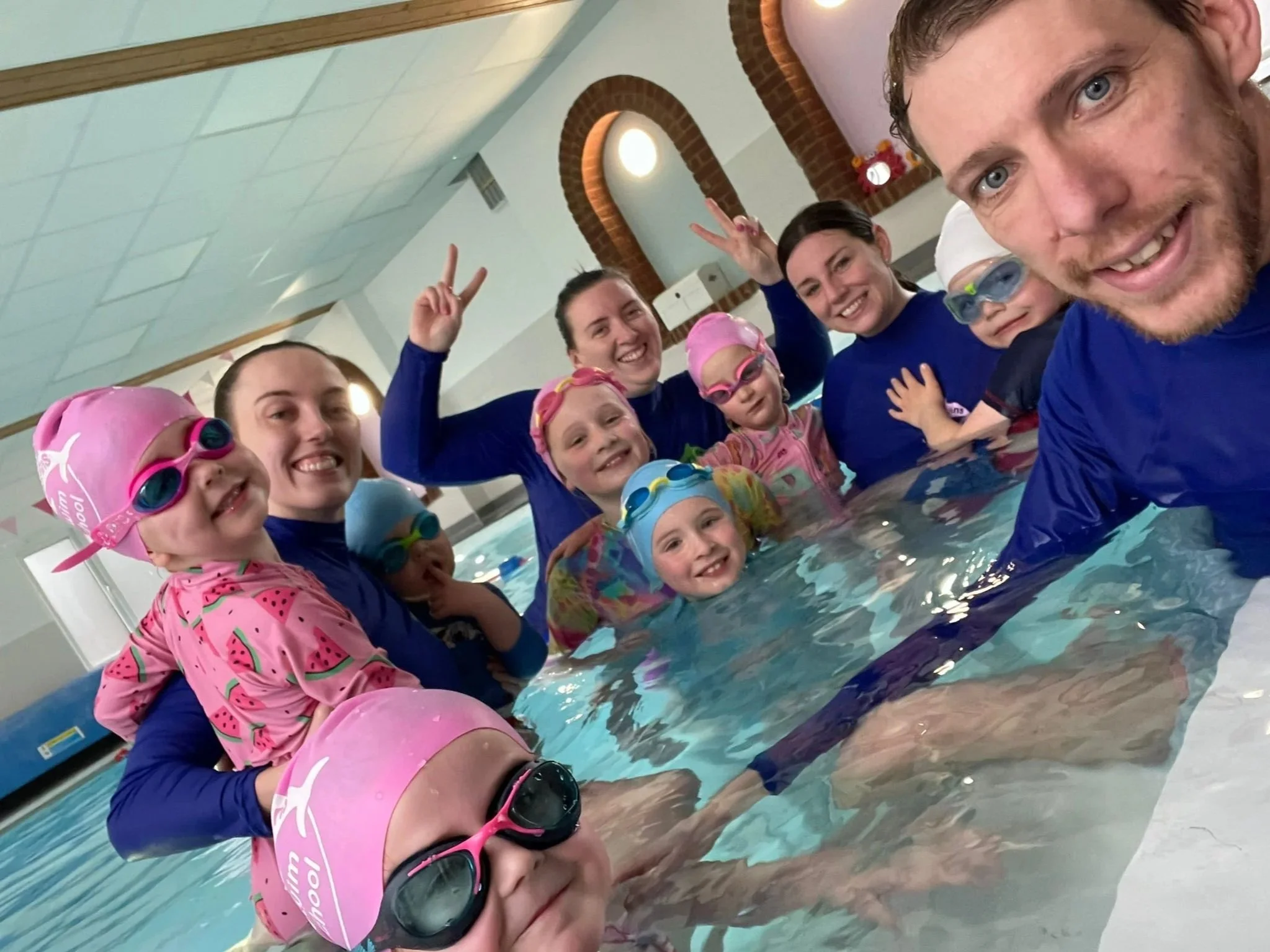 Happy Fins teachers and young swimmers squeeze into a smiling group selfie in the pool