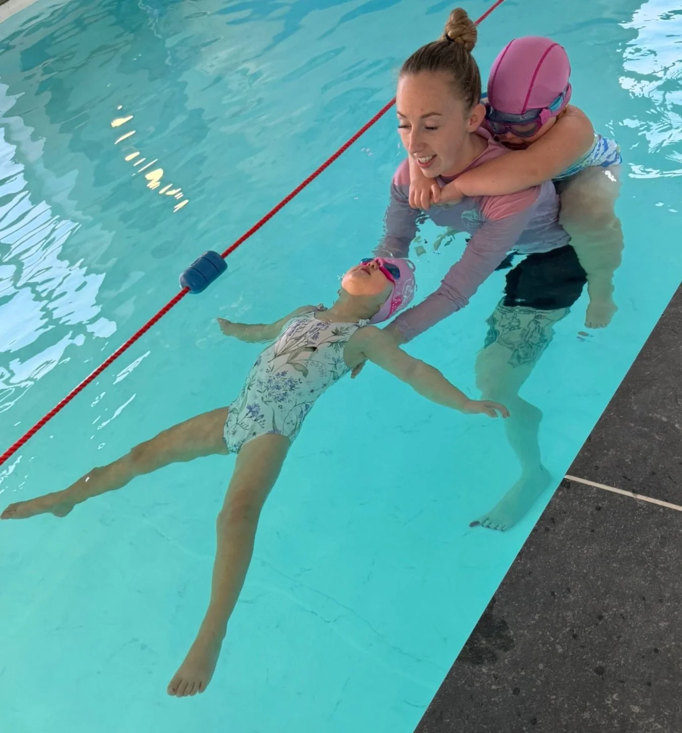 A Happy Fins teacher helps one young swimmer float while another holds on to her back