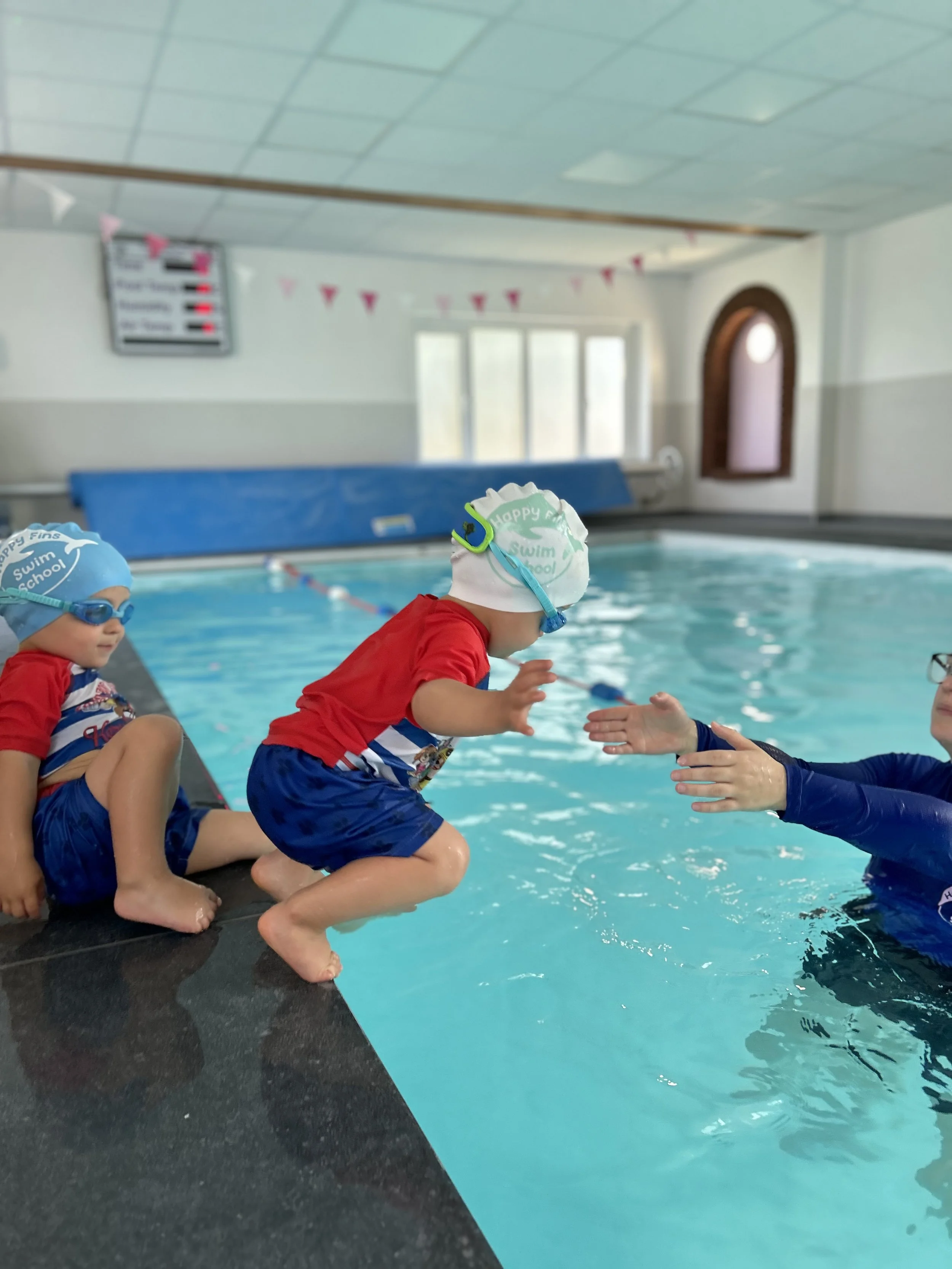 A young swimmer steps off the pool edge towards their teacher's outstretched hands, with no armbands