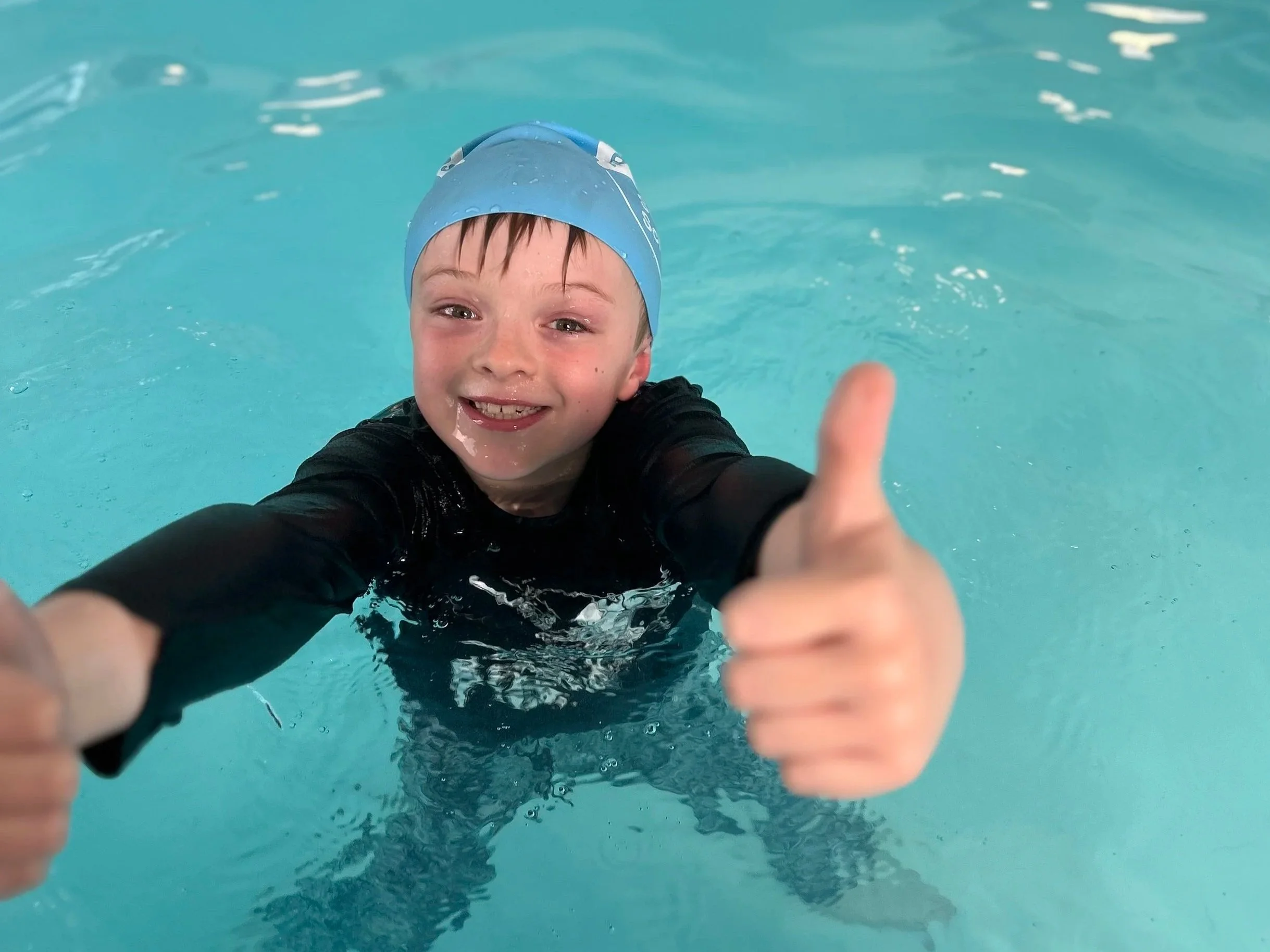 A young swimmer in a swim cap gives a big thumbs up in the water