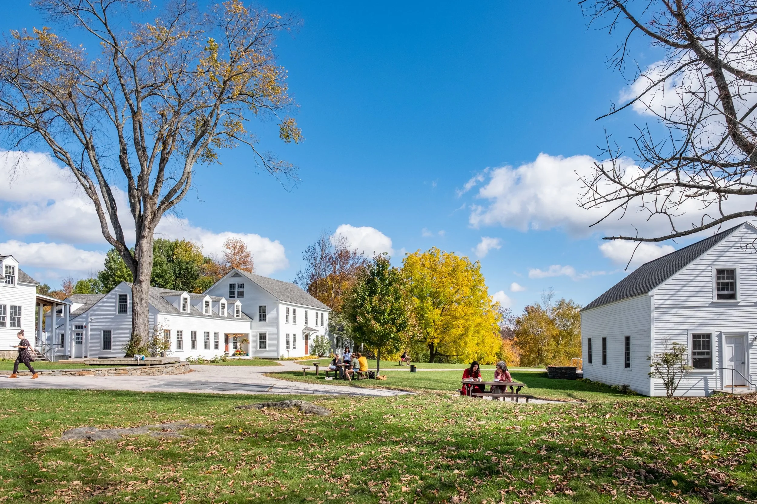 A park scene with several white houses, green grass, trees with colorful autumn leaves, and people sitting at picnic tables under a bright blue sky with white clouds.
