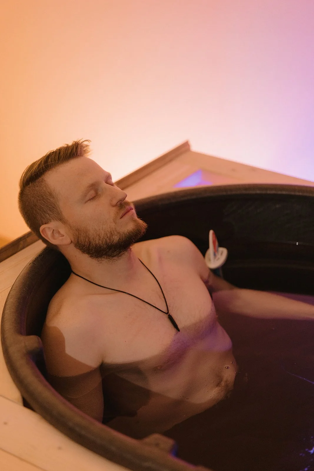 A man with closed eyes relaxing in a wooden hot tub filled with water, wearing a black necklace with a pendant, with a peaceful expression on his face.