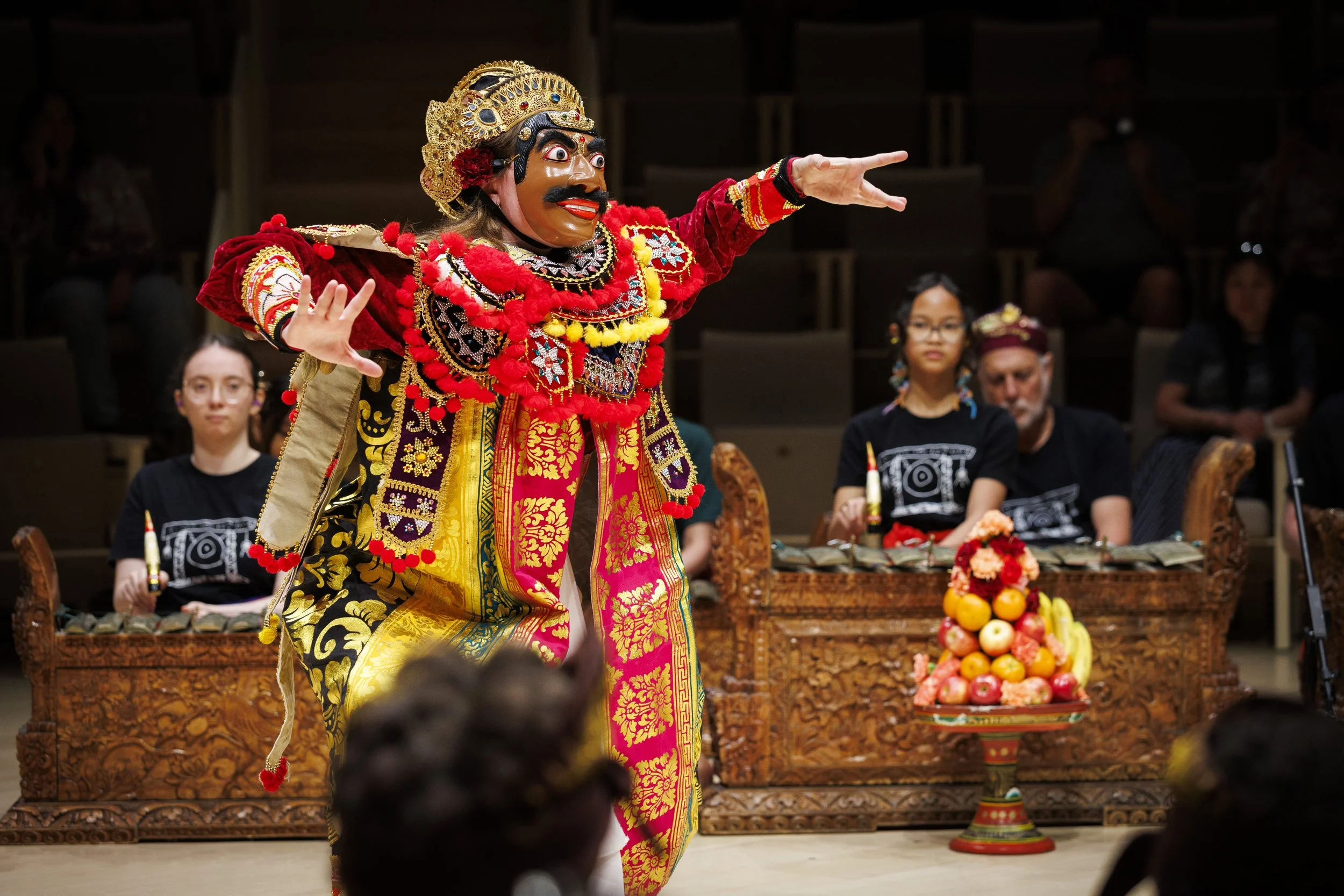 A performer in traditional costume and mask dances in front of an audience at a cultural event, with a basket of fruit and flowers in the background.