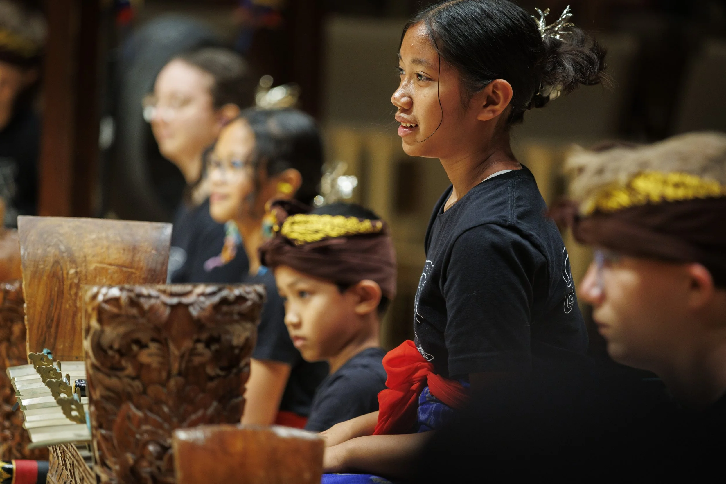 A group of children and a woman, possibly a teacher, sitting at a table with traditional decorative items in front of them, in an indoor setting.