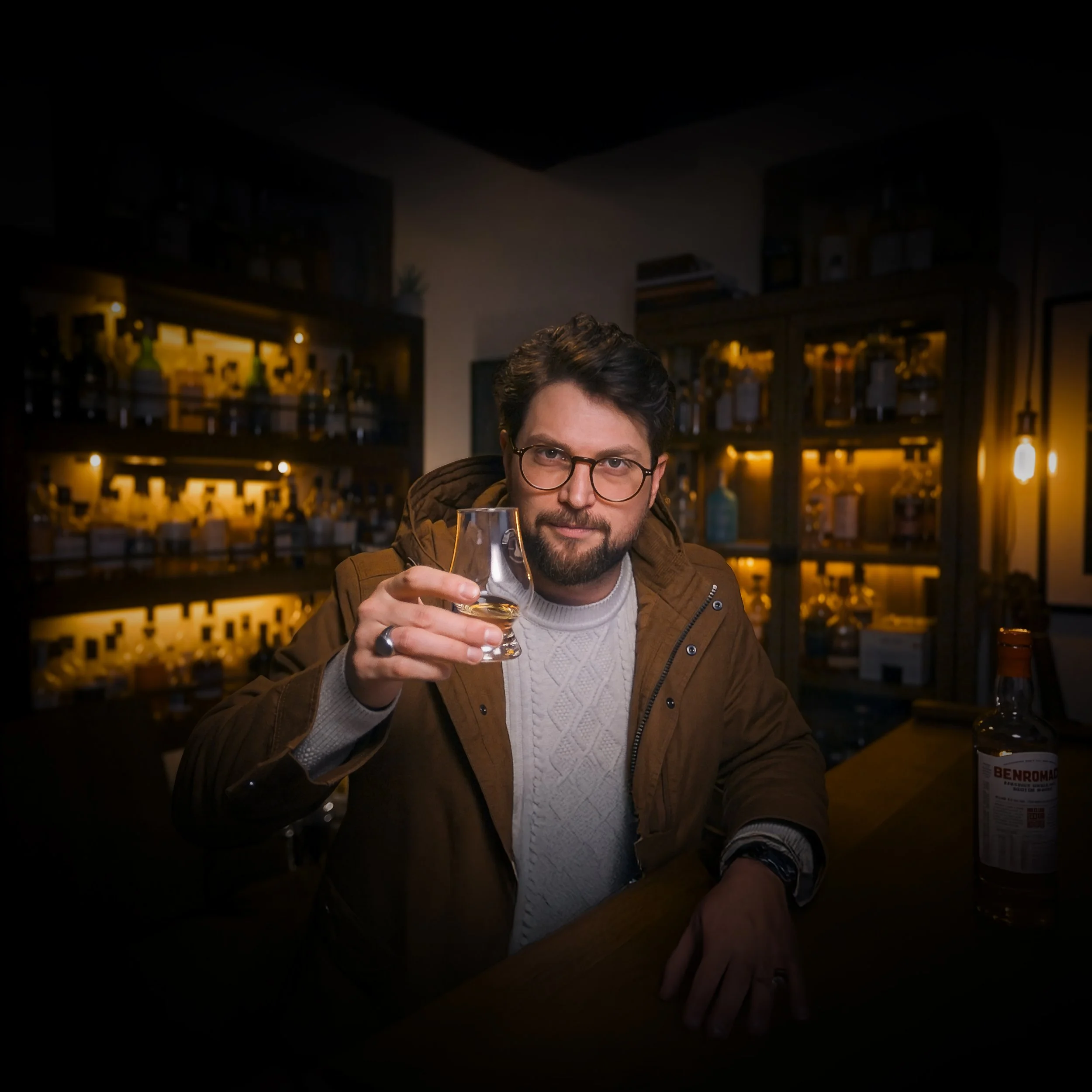 A man with glasses and a beard sitting at a bar holding a glass of whiskey, with shelves of liquor bottles behind him.