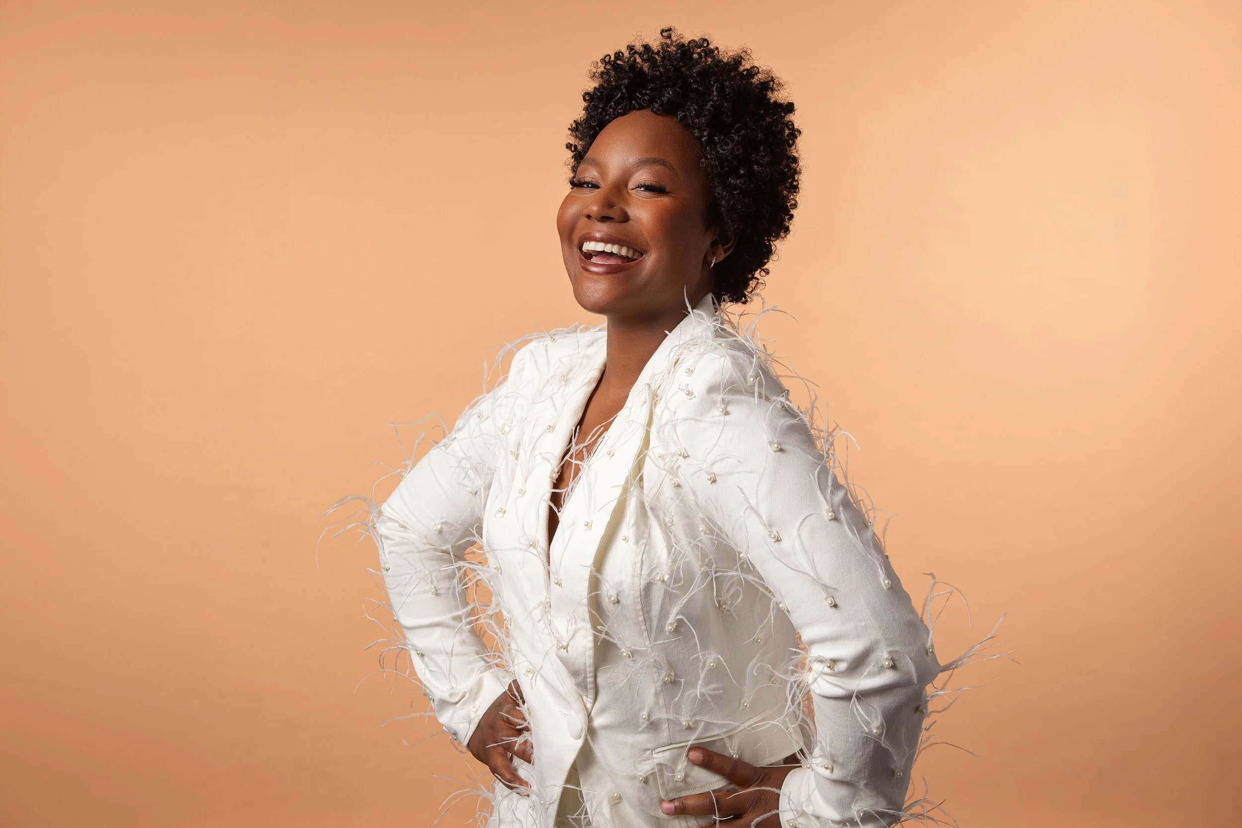 A woman with dark skin and short, curly black hair smiling confidently while wearing a white jacket with feather-like embellishments and pearls, against a peach-colored background.