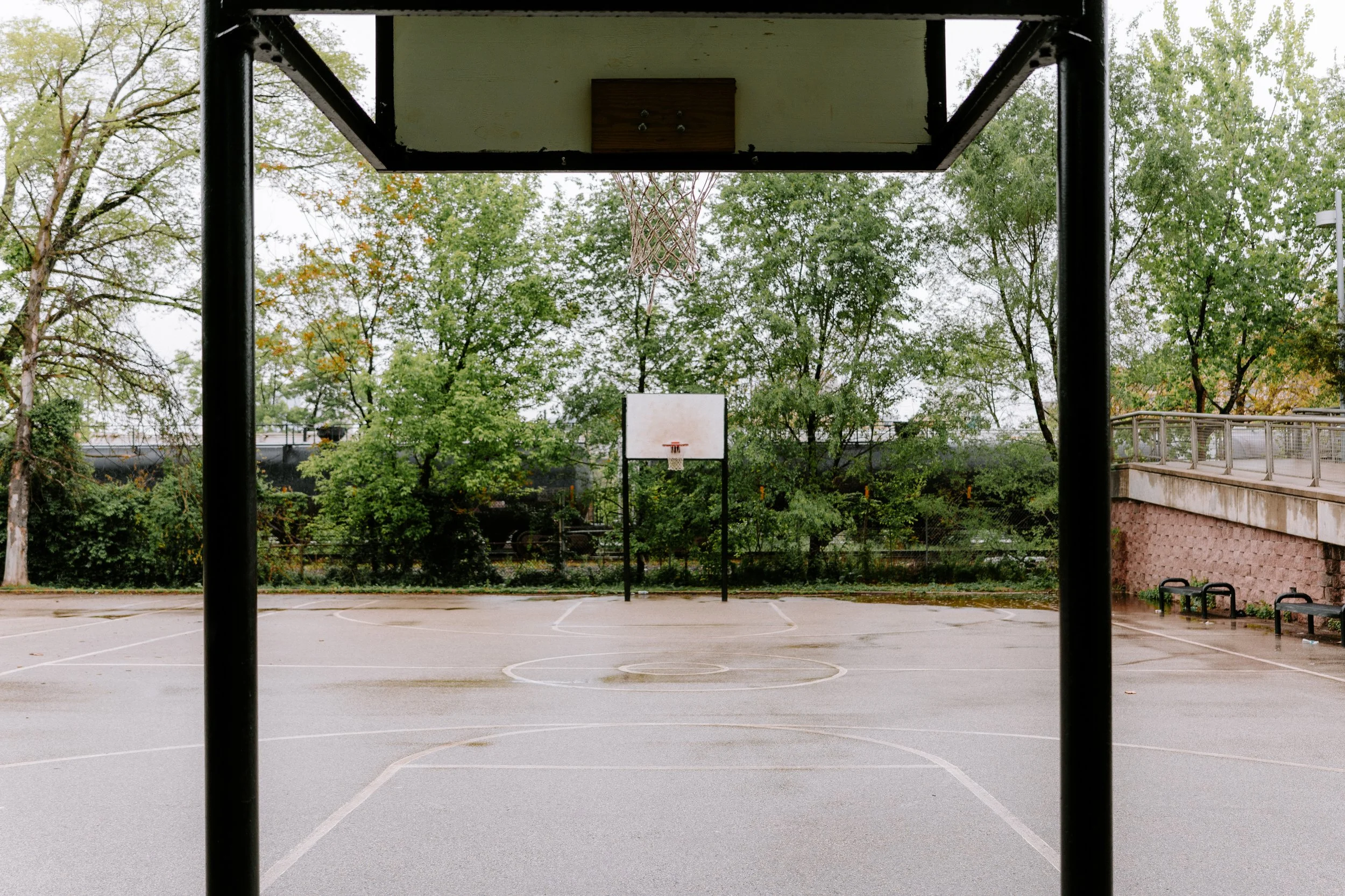 Empty outdoor basketball court viewed from under the basketball hoop, with trees and a fence in the background.