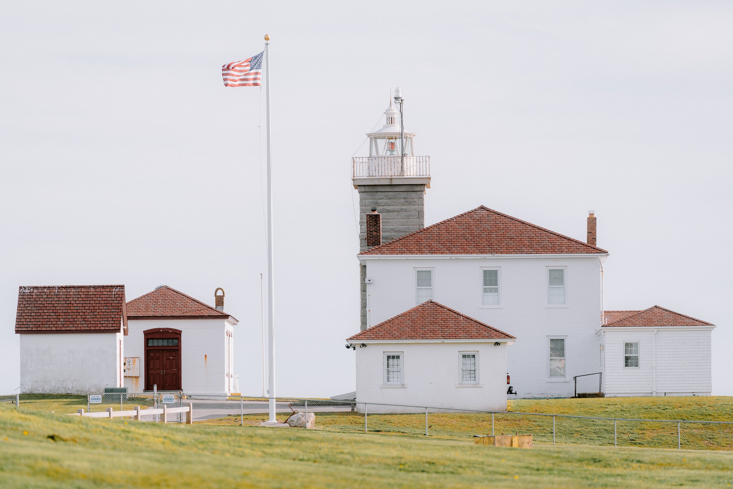 A lighthouse with an American flag flying on a tall flagpole, white buildings with red roofs, surrounded by a grassy area with a fence.
