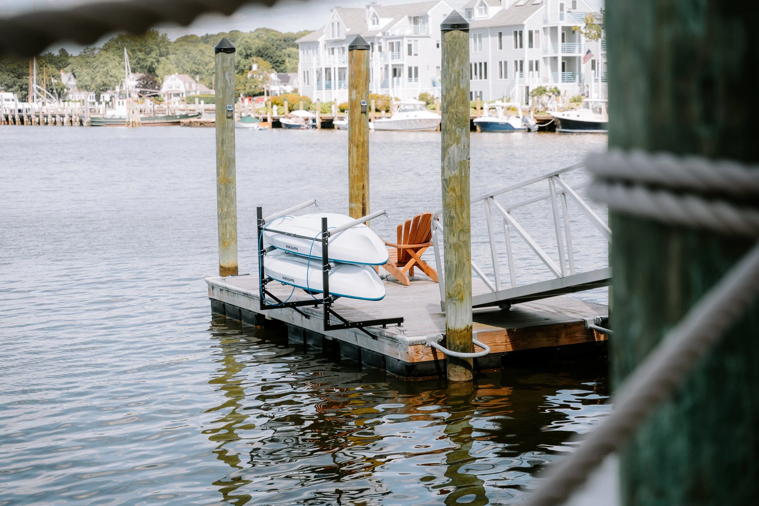 A marina dock with two stacked kayaks, a wooden Adirondack chair, and a ramp leading to a boat storage area, with houses and boats in the background.
