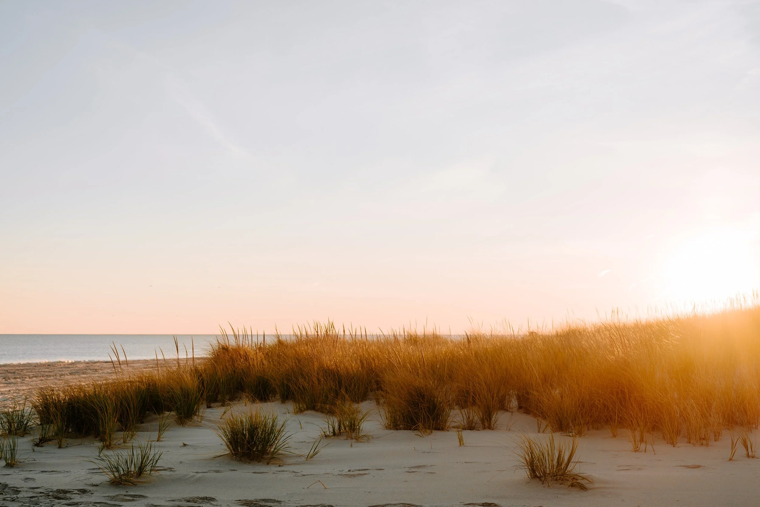 Beach scene at sunset with sandy dunes and grass, calm ocean in the background, and the sun near the horizon.