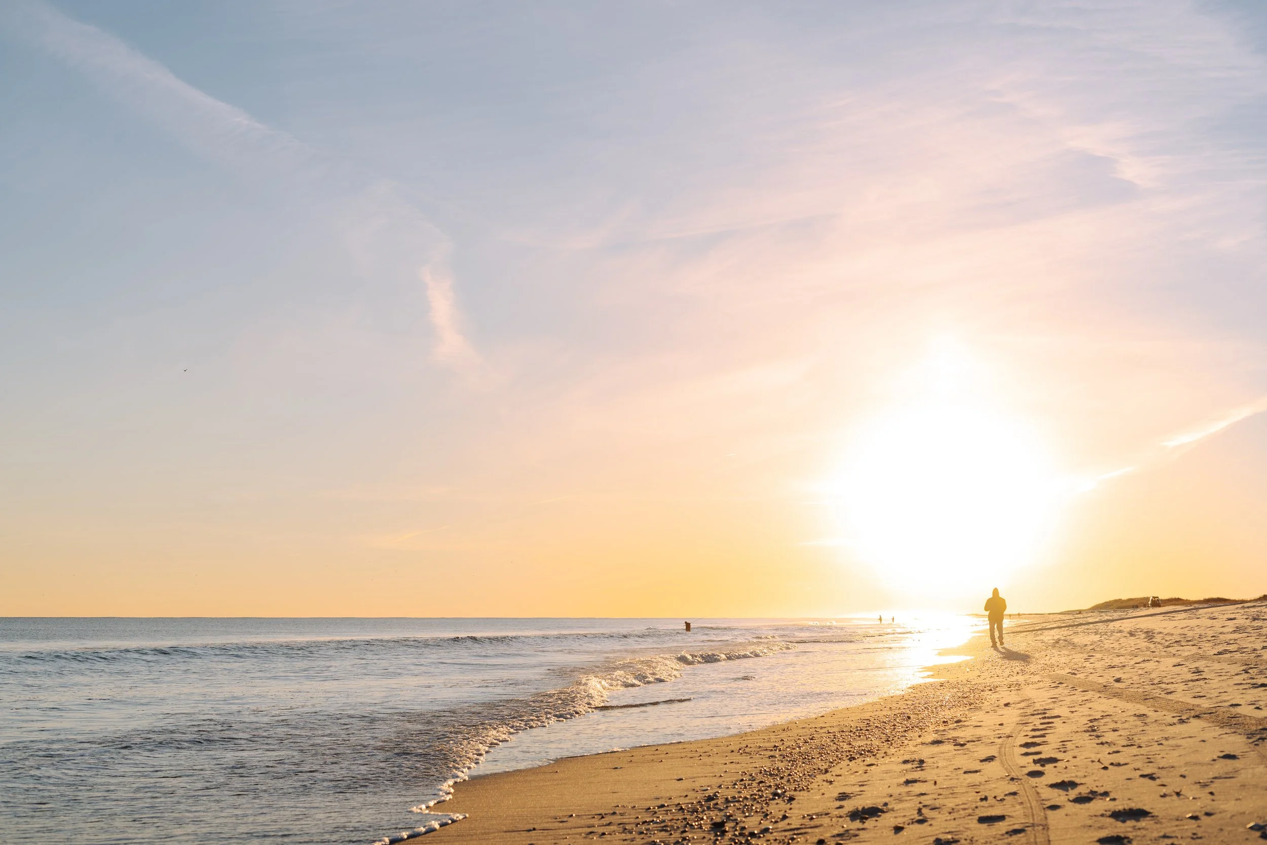A person walking along the beach at sunset with the sun low on the horizon, casting a bright glow and creating long shadows on the sand.