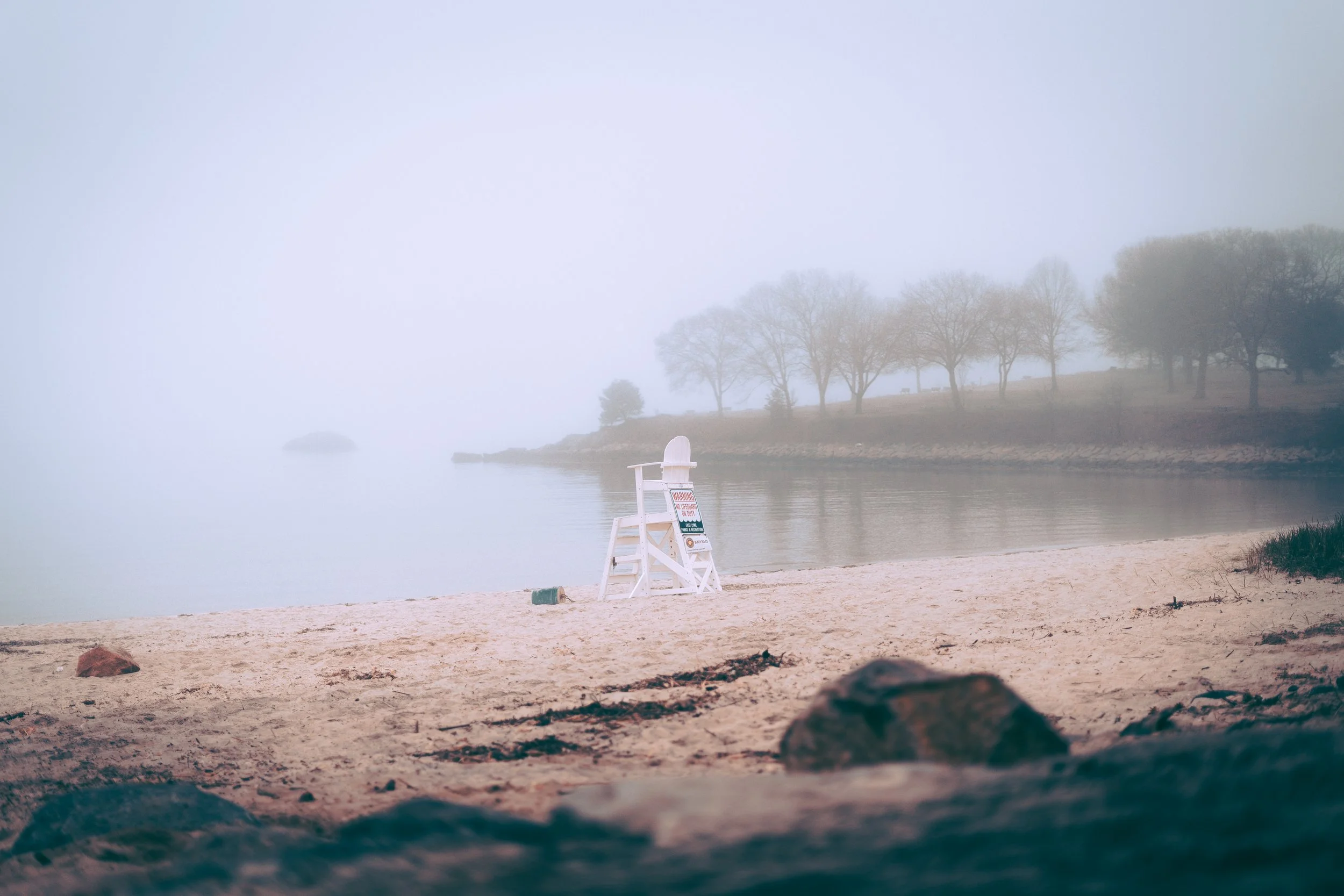 A foggy lakeside beach with a lifeguard chair and a ‘Warning’ sign, surrounded by rocks and trees in the background.