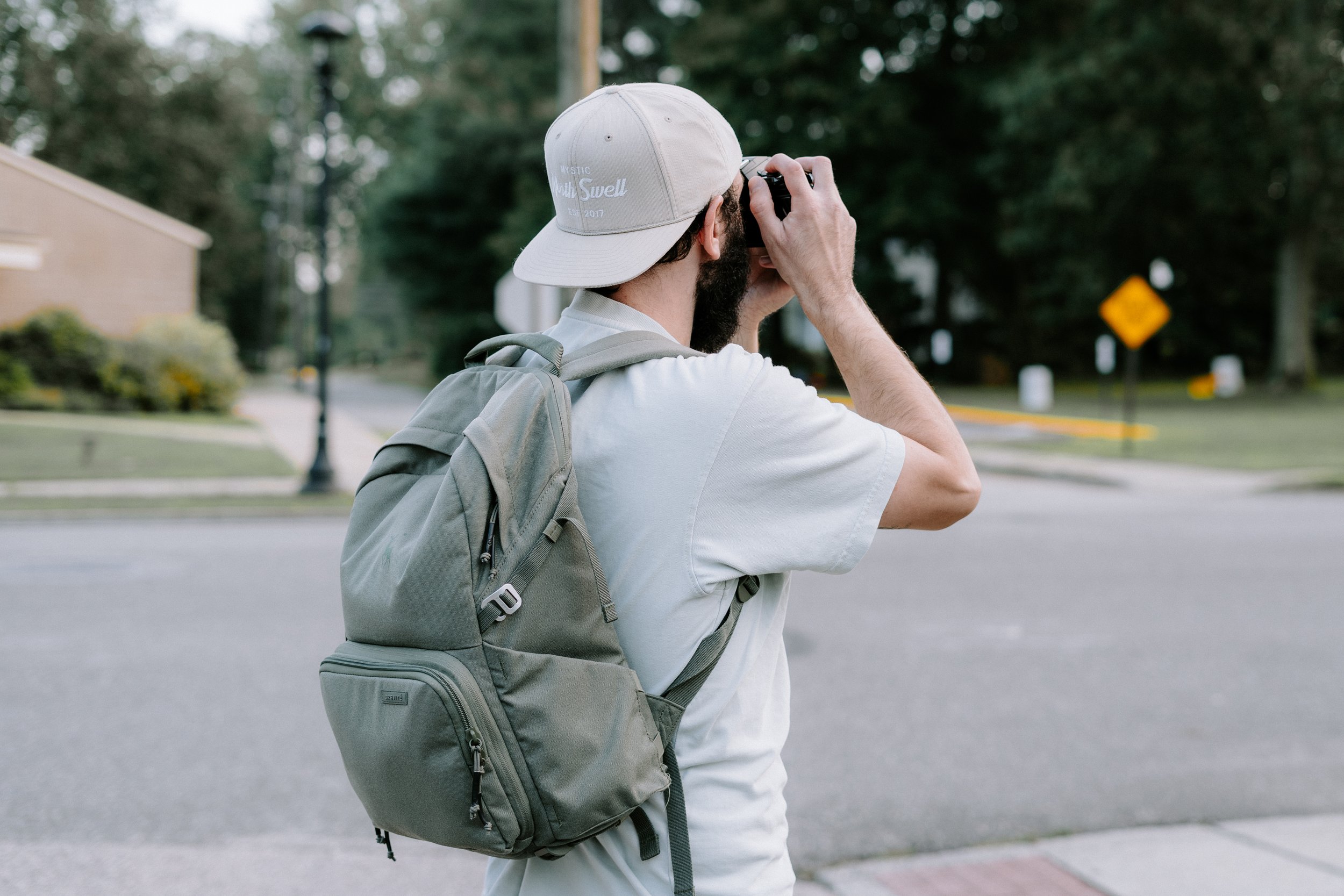A man with a backpack takes a photograph with a camera on a street in a suburban neighborhood with trees and houses in the background.