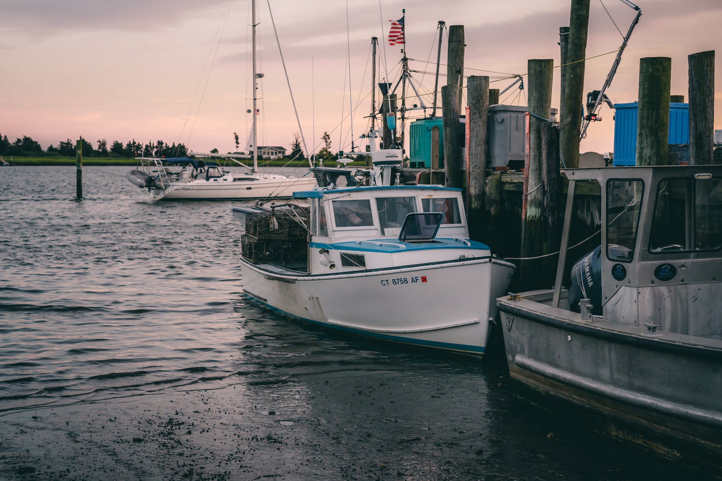 Marina with boats docked at the pier during sunset, including a small white fishing boat with a blue stripe in the foreground and sailboats in the background.