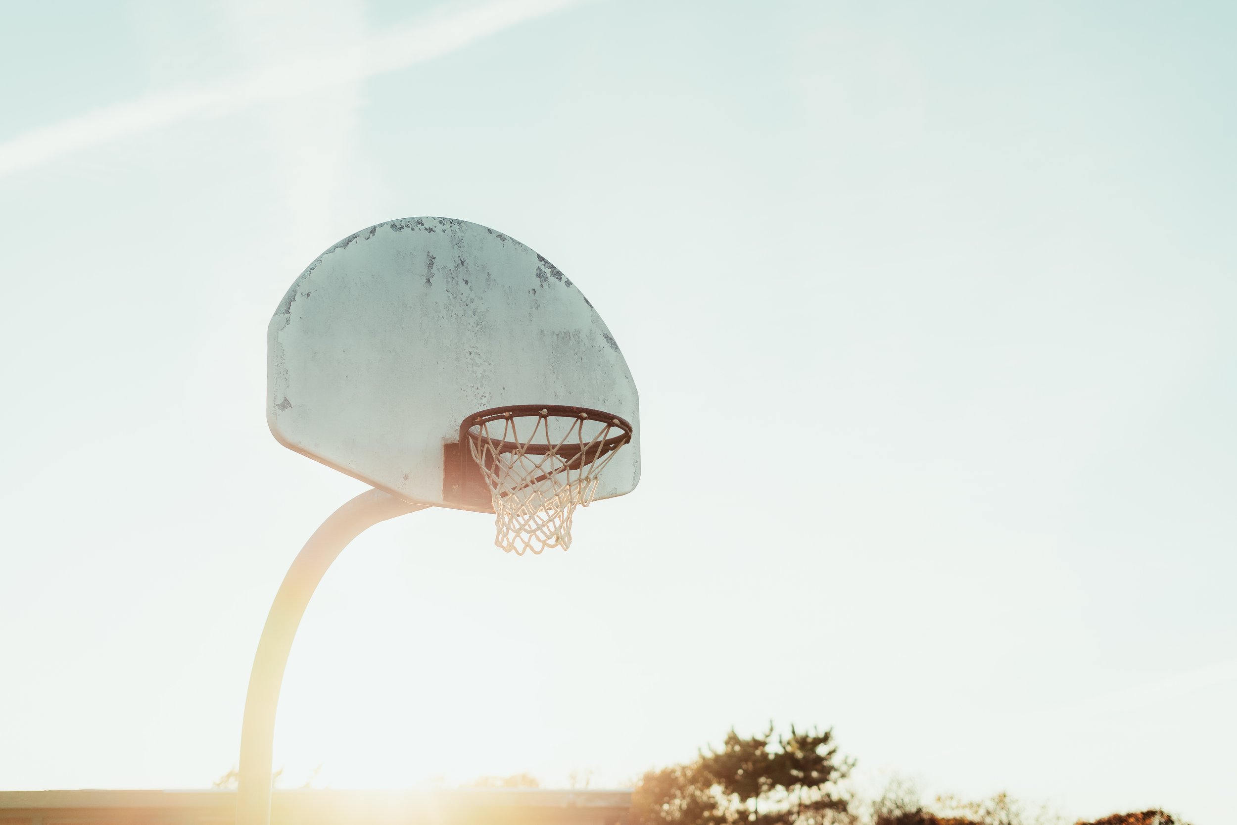An outdoor basketball hoop with a weathered backboard and net, set against a bright sky with trees in the background.