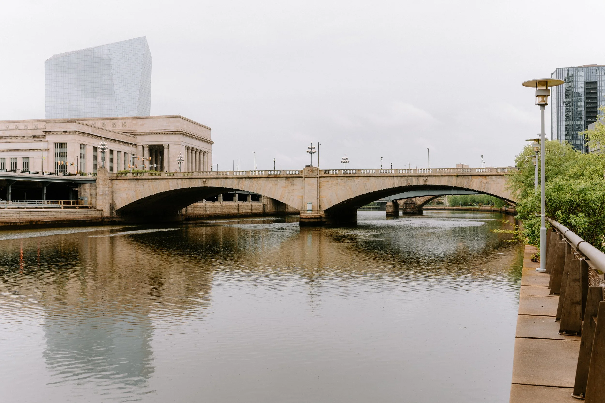Cityscape with a stone bridge over a river, modern buildings in the background, trees, and street lamps along a riverwalk.