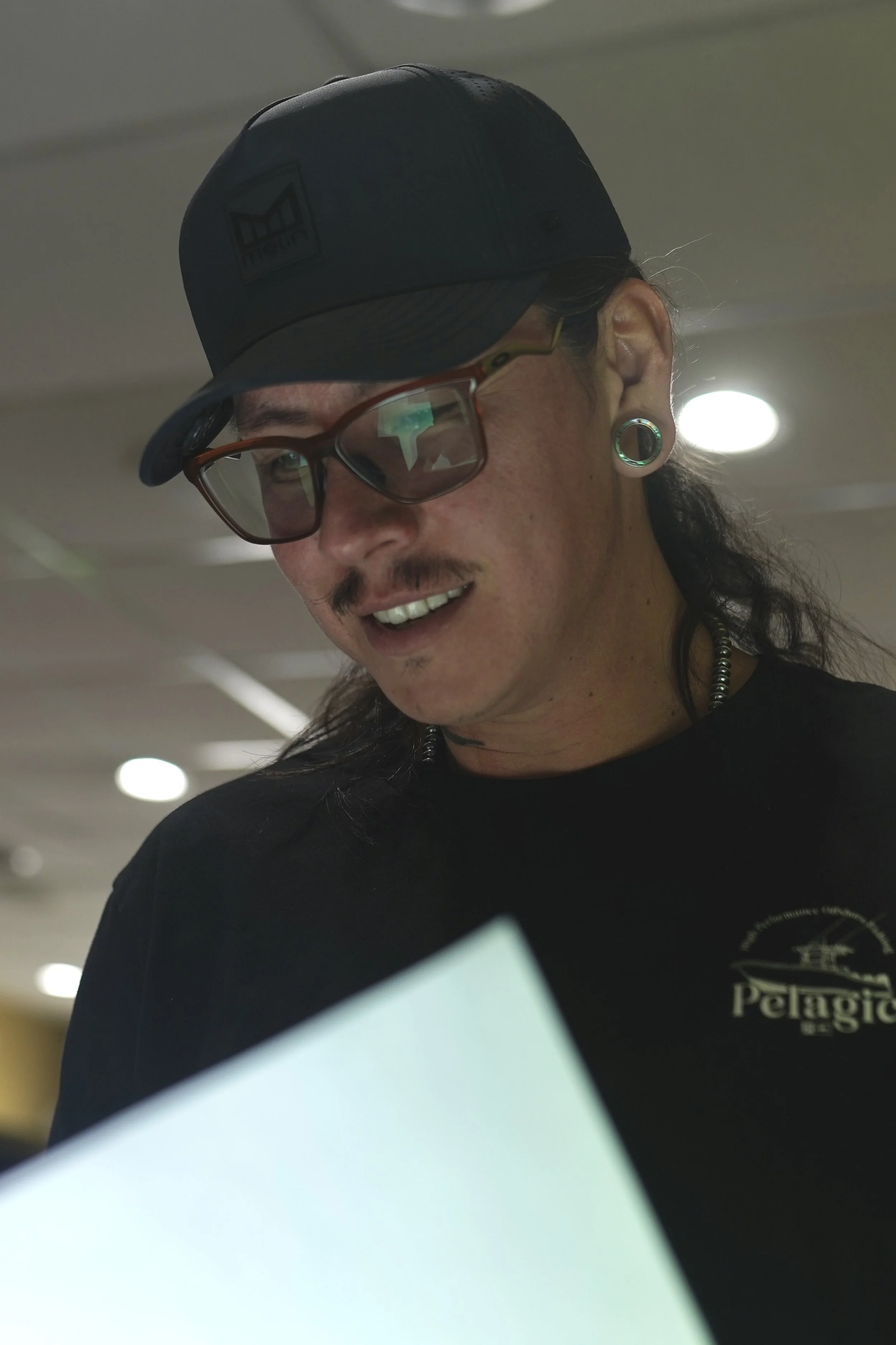 A man wearing glasses, a black baseball cap, and black shirt with a logo, smiling and looking downward in an indoor setting with bright lights.