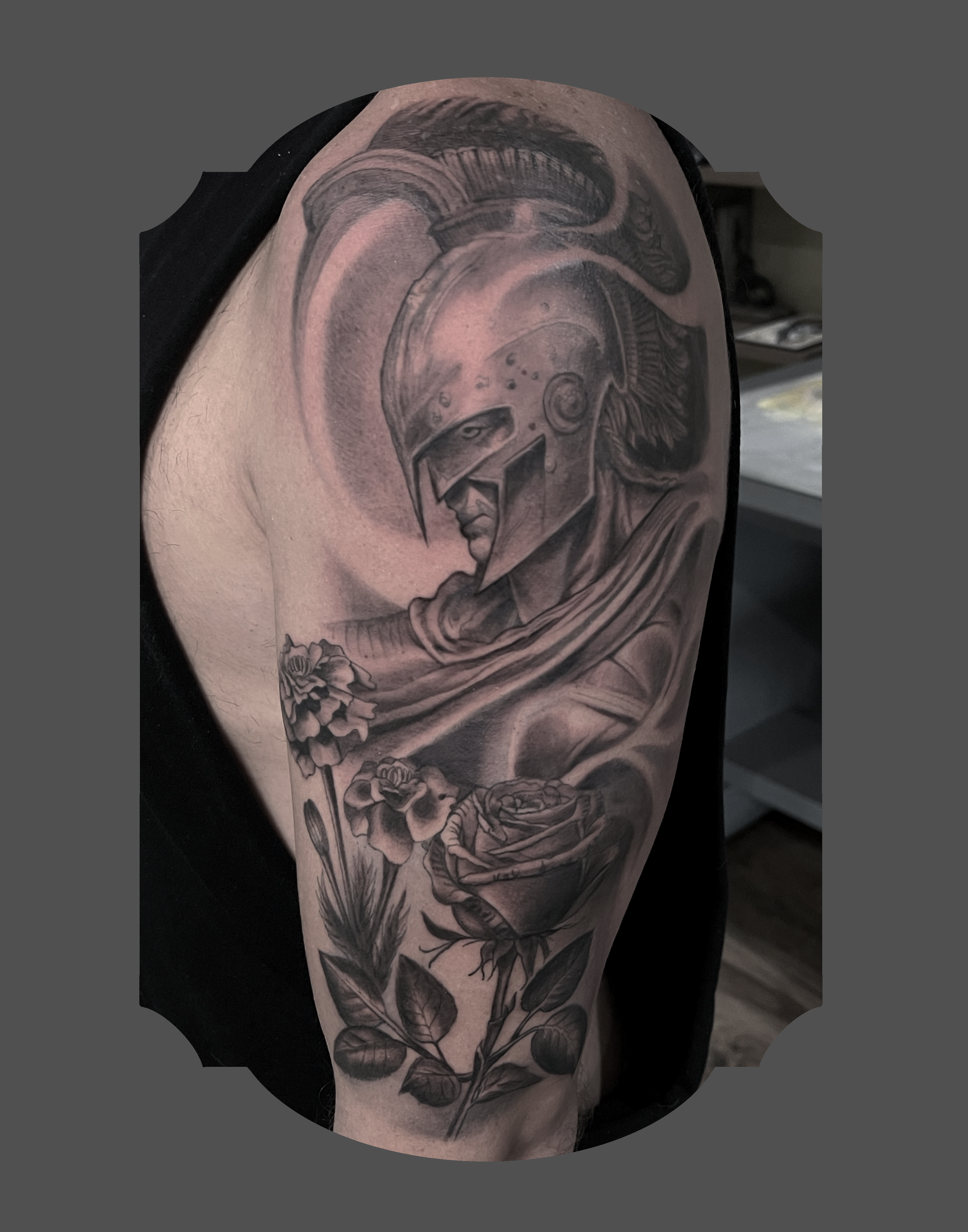 Black and grey tattoo on upper arm depicting a Spartan warrior helmet with detailed shading, roses, and leaves.