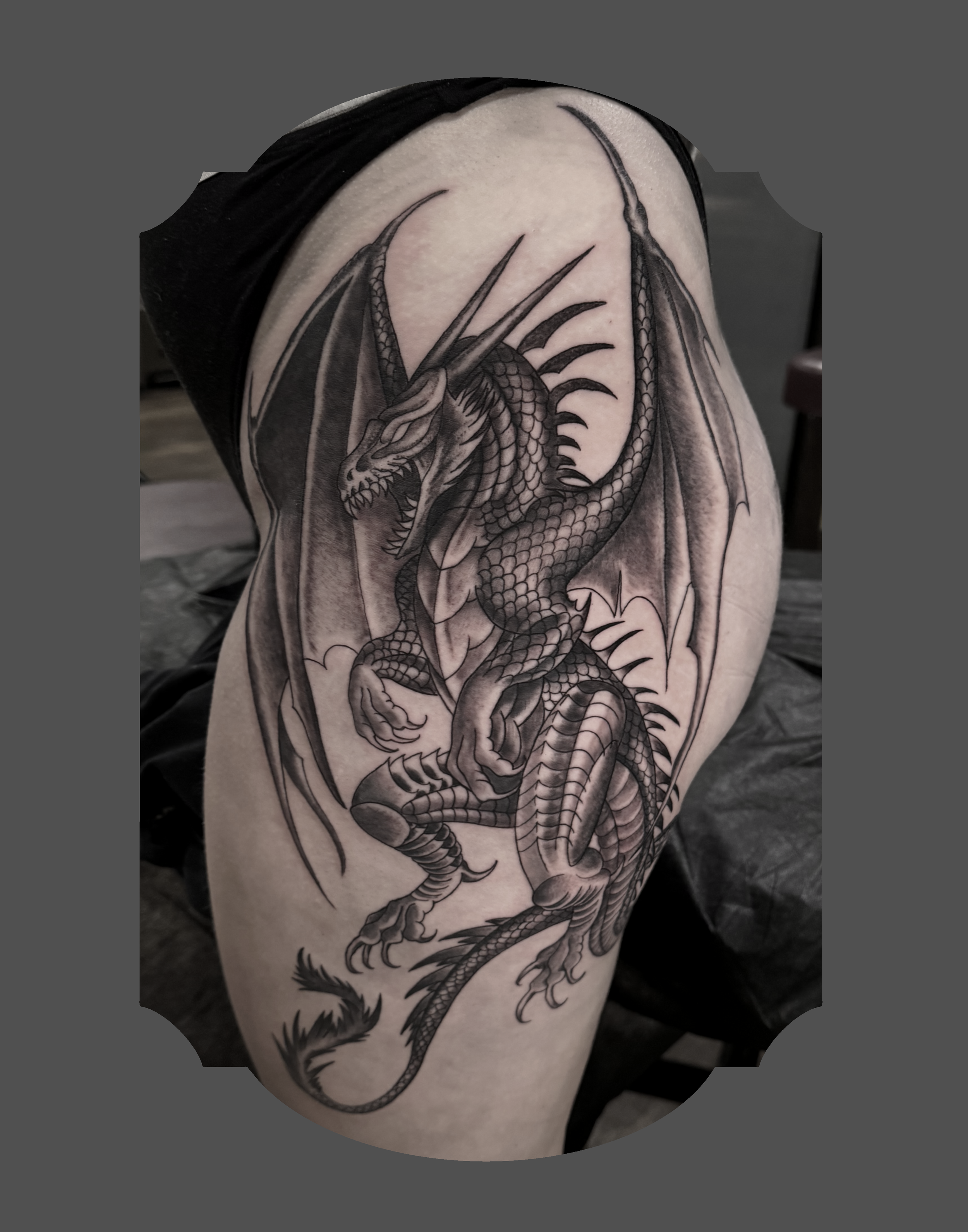 Black and gray tattoo of a fierce dragon with detailed scales, wings, and claws on a person's upper arm.