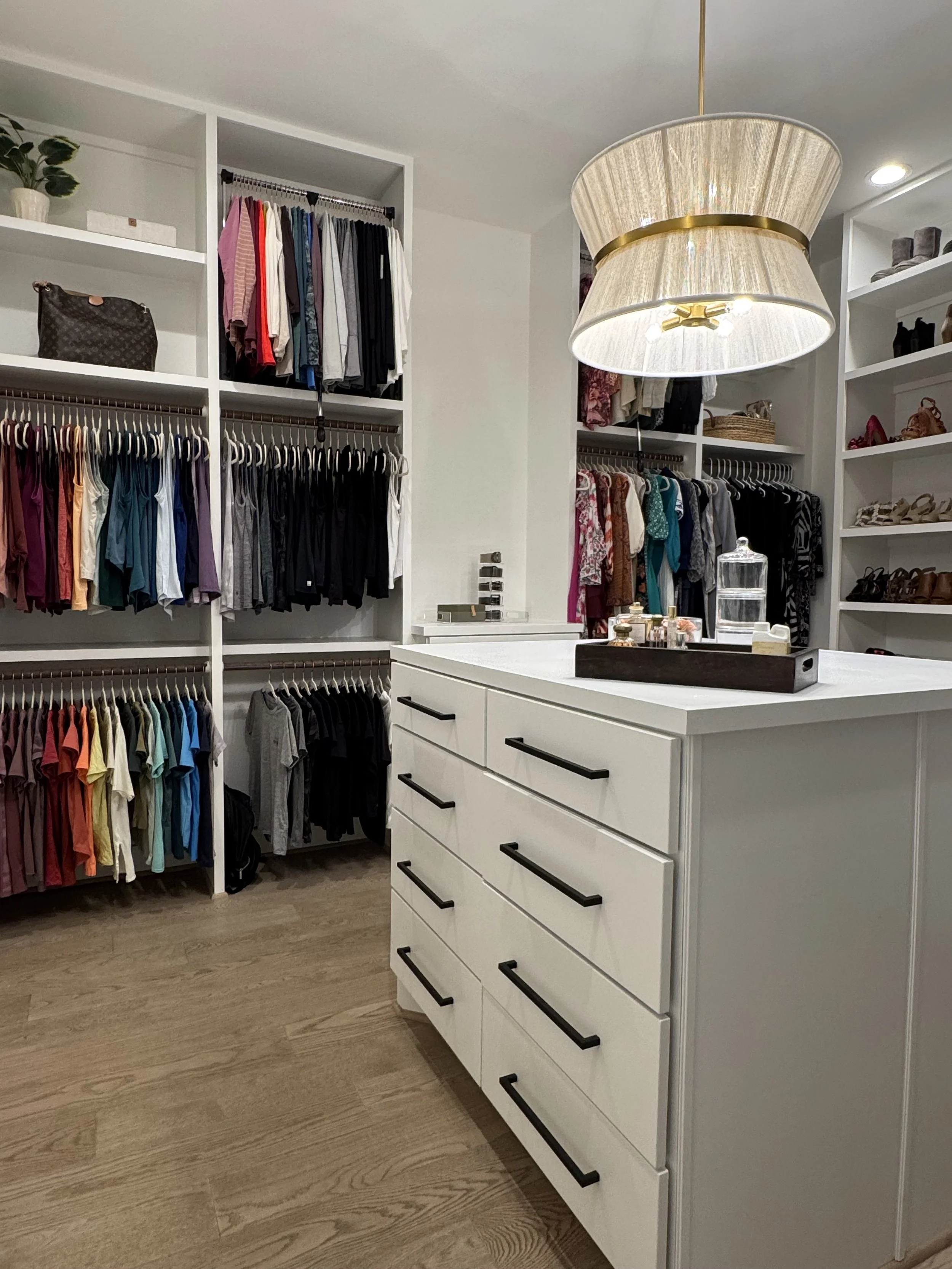 A walk-in closet with white shelves and drawers filled with colorful clothes, shoes, and accessories, illuminated by a large hanging light fixture.