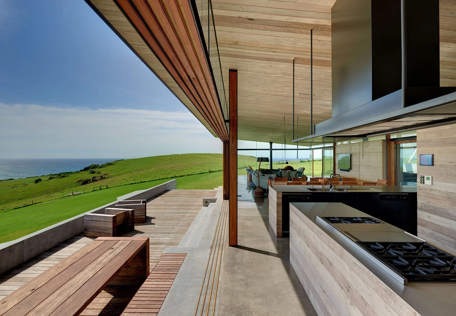 Modern house with open kitchen and living area, large glass windows, wooden ceiling, and outdoor patio overlooking green hills and ocean.