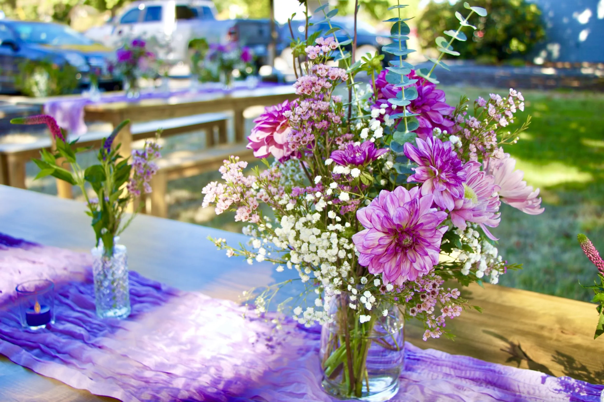 Outdoor picnic setup with a low wooden table decorated with pink and white flowers, purple candles, and elegant tableware on a lace table runner, surrounded by cushions and pillows on a grassy lawn.