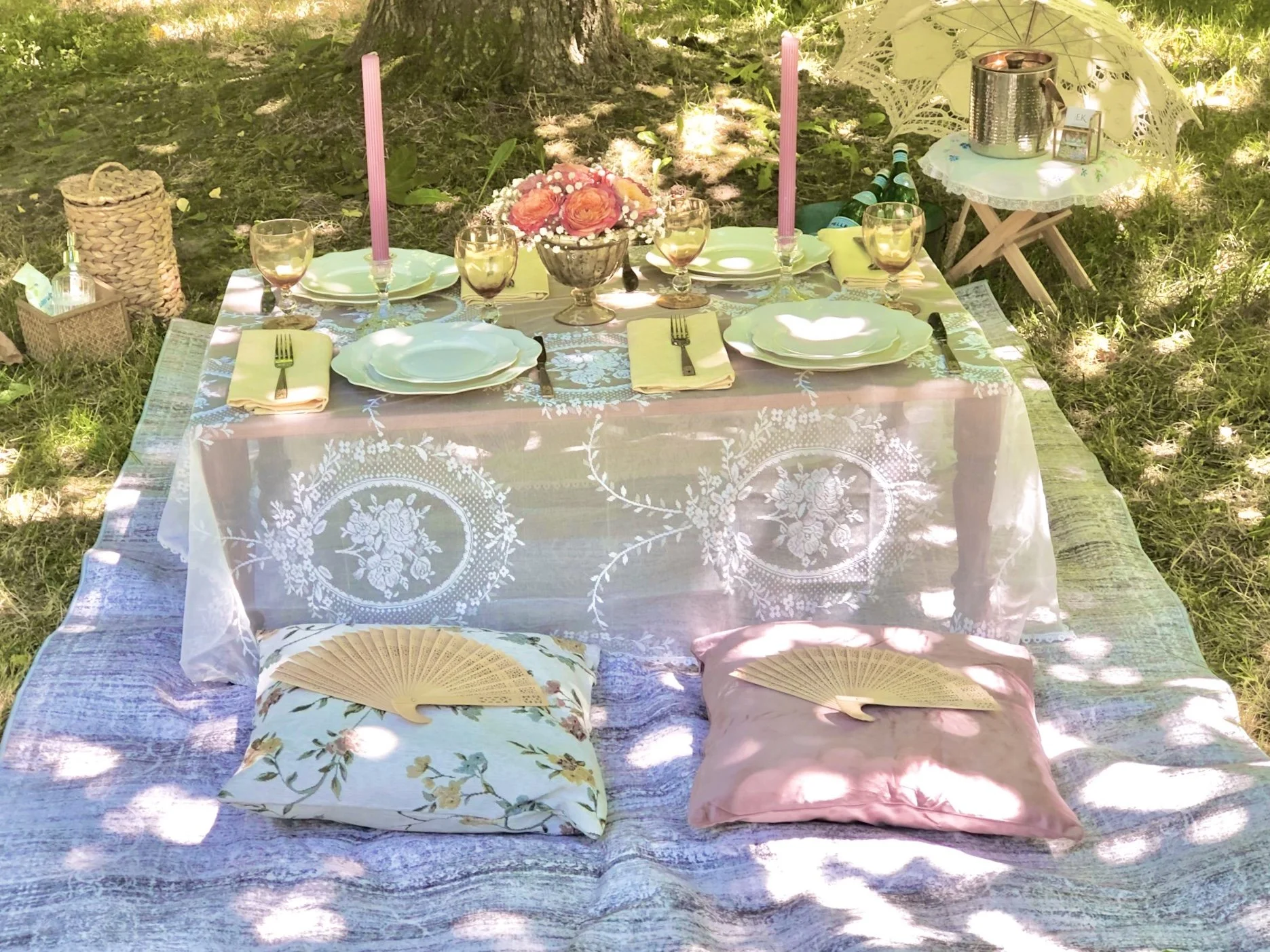 Elegant outdoor dining table set for a picnic, with white tablecloth, floral arrangements, candlesticks, black stemware, gold-rimmed plates, and place settings, situated on a grassy lawn with cushions and decorative items in the background.