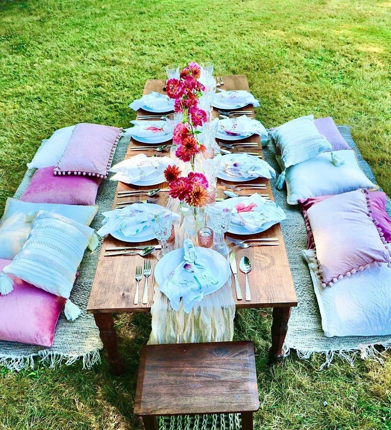 A formal outdoor dinner setup on a grass lawn with a low table covered in a cream tablecloth, surrounded by cushions and pillows for seating. The table is decorated with blue and white plates, blue napkins, glassware, and candles, with a centerpiece of white and pink flowers in a white vase. A small side table with a jug, bottle, and a letter boardis also visible.