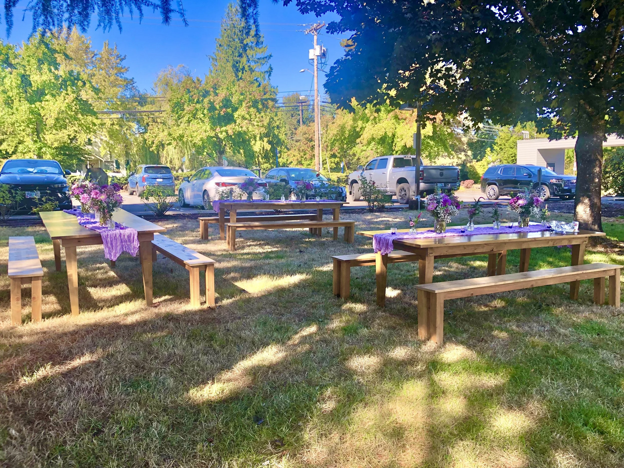 Elegant outdoor dining table set for a picnic, with white tablecloth, floral arrangements, candlesticks, black stemware, gold-rimmed plates, and place settings, situated on a grassy lawn with cushions and decorative items in the background.