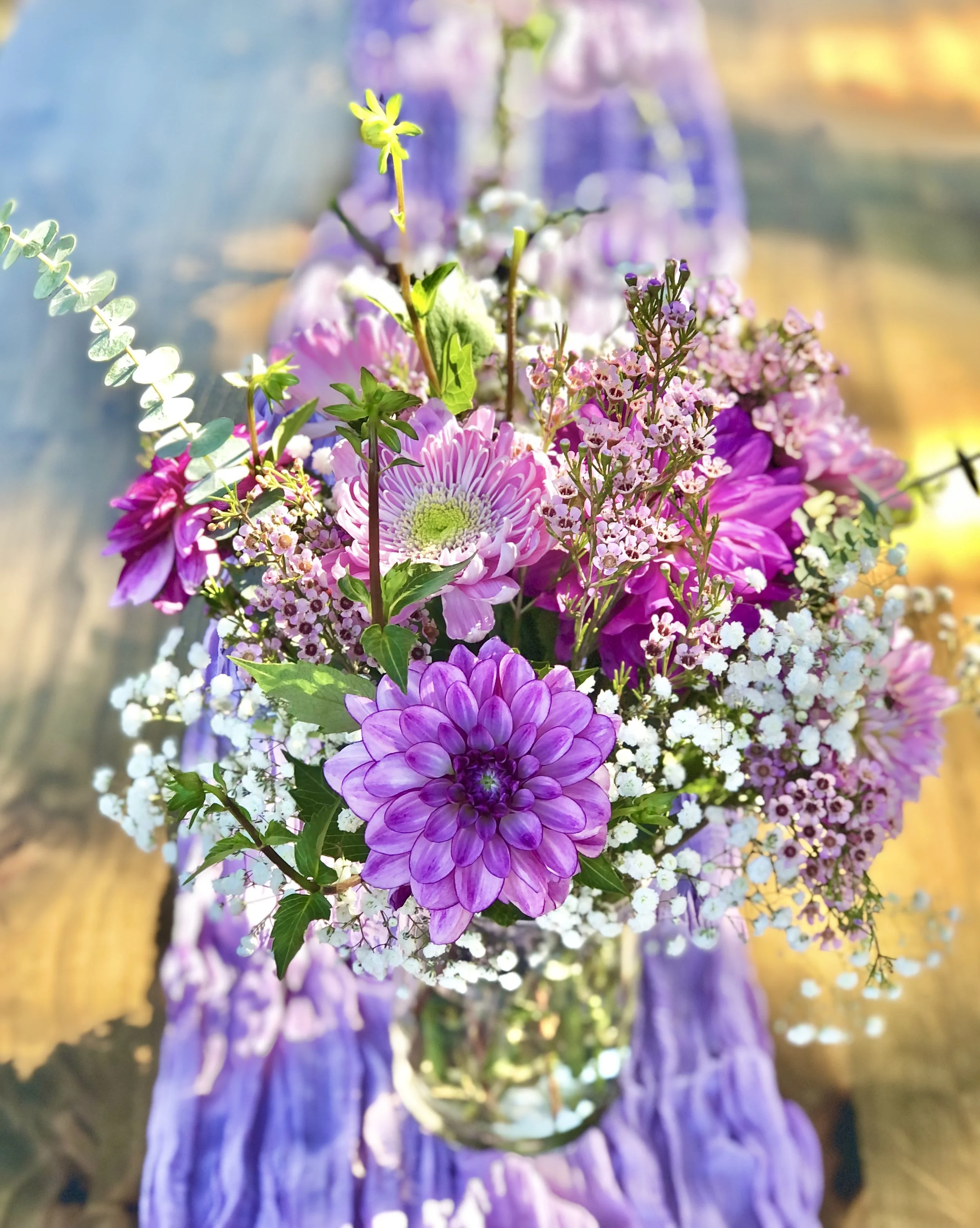 A picnic setup on the grass with a table covered in a floral tablecloth, decorated with candles, glasses, plates, and a large white vase with purple flowers. Next to the table is a woven rug with pillows, and a basket of food and grapes placed on it.