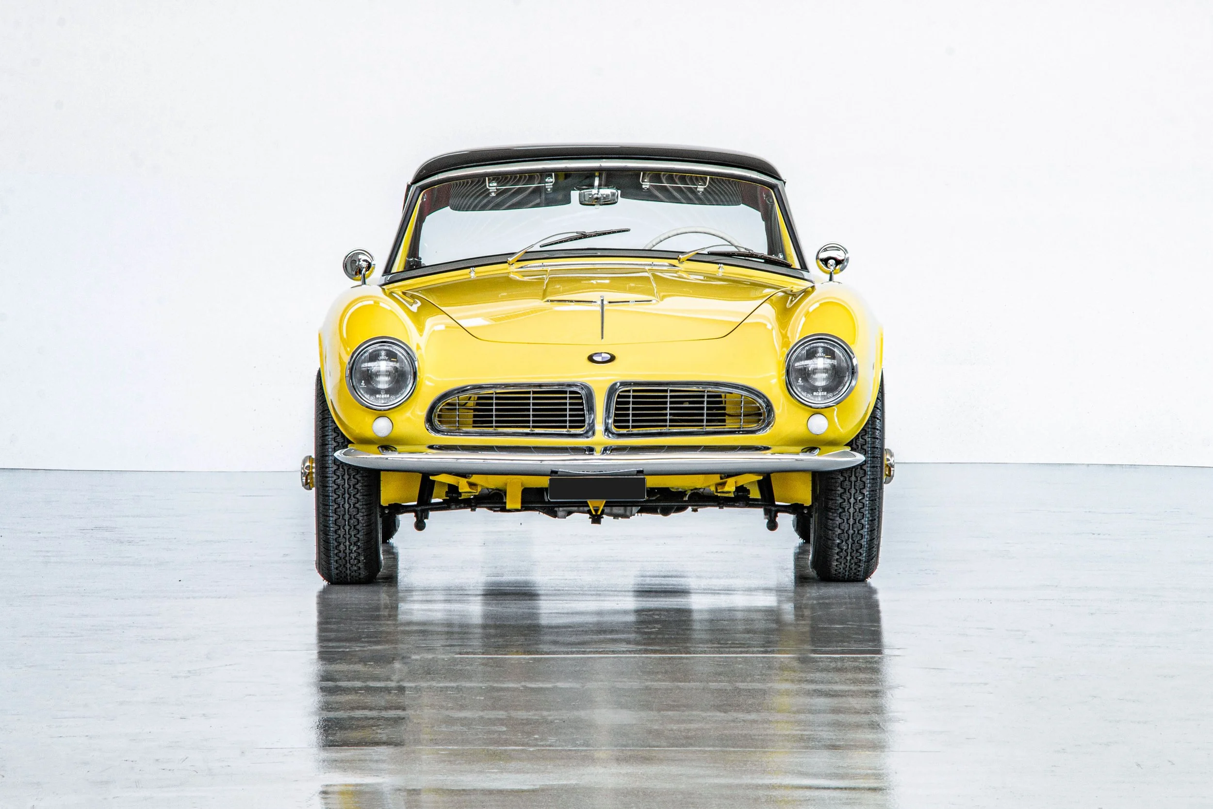 Front view of a vintage yellow car with black roof parked indoors on a reflective floor against a white wall.