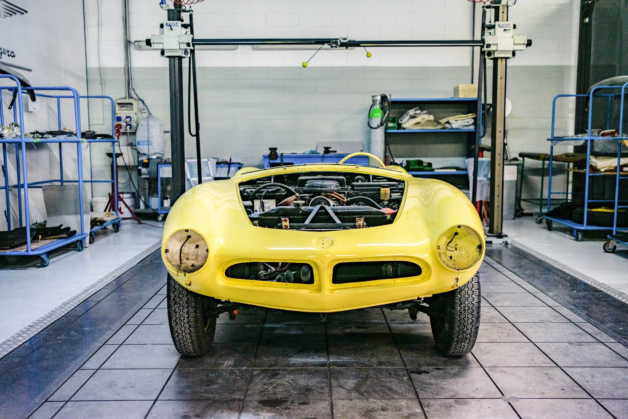 A yellow vintage race car with its hood removed, showing the engine, is on a vehicle lift in a garage or workshop with tools and shelves in the background.