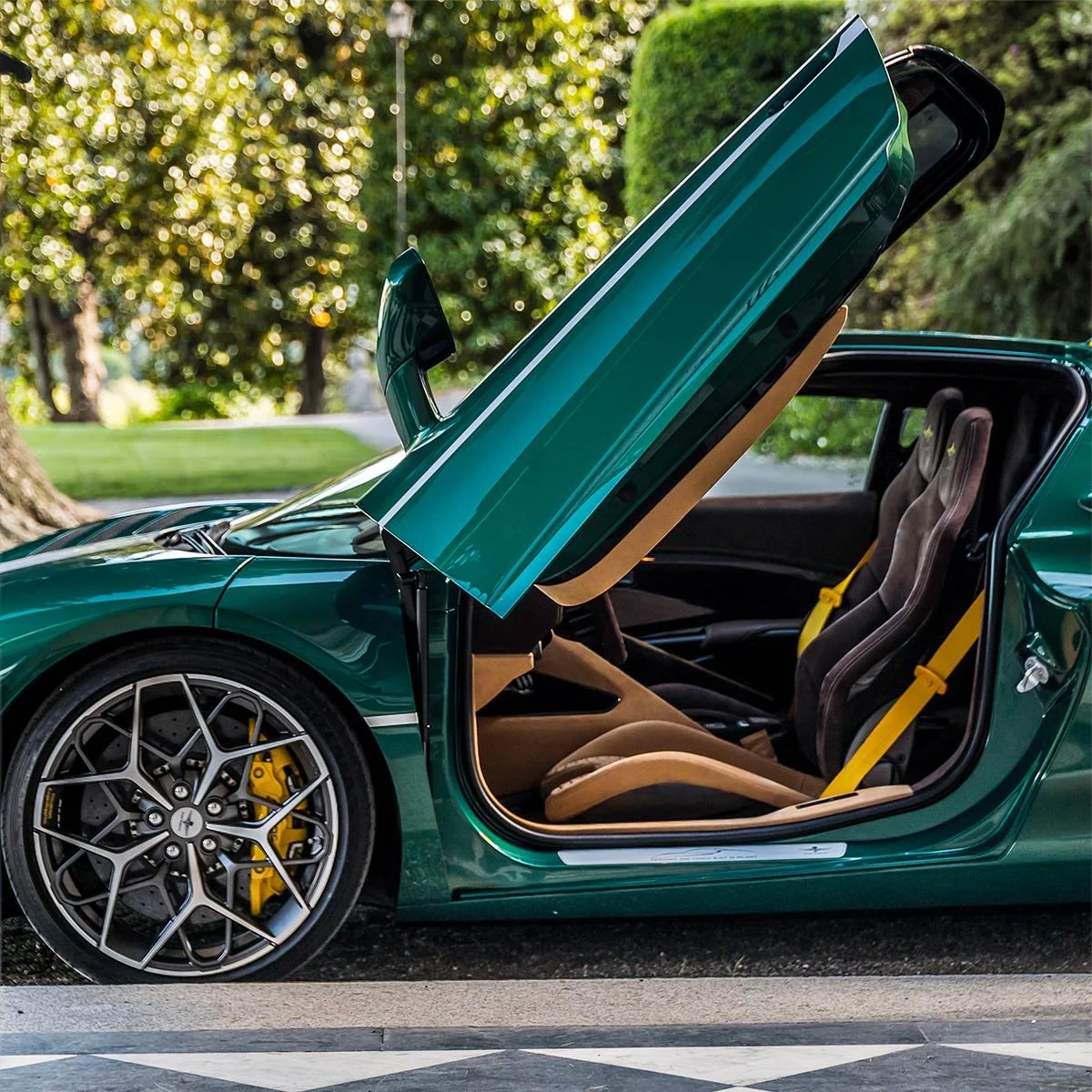 A green sports car with scissor doors open, revealing black racing seats with yellow seatbelts and yellow brake calipers behind stylish multi-spoke wheels, parked on a street with green trees and grass in the background.