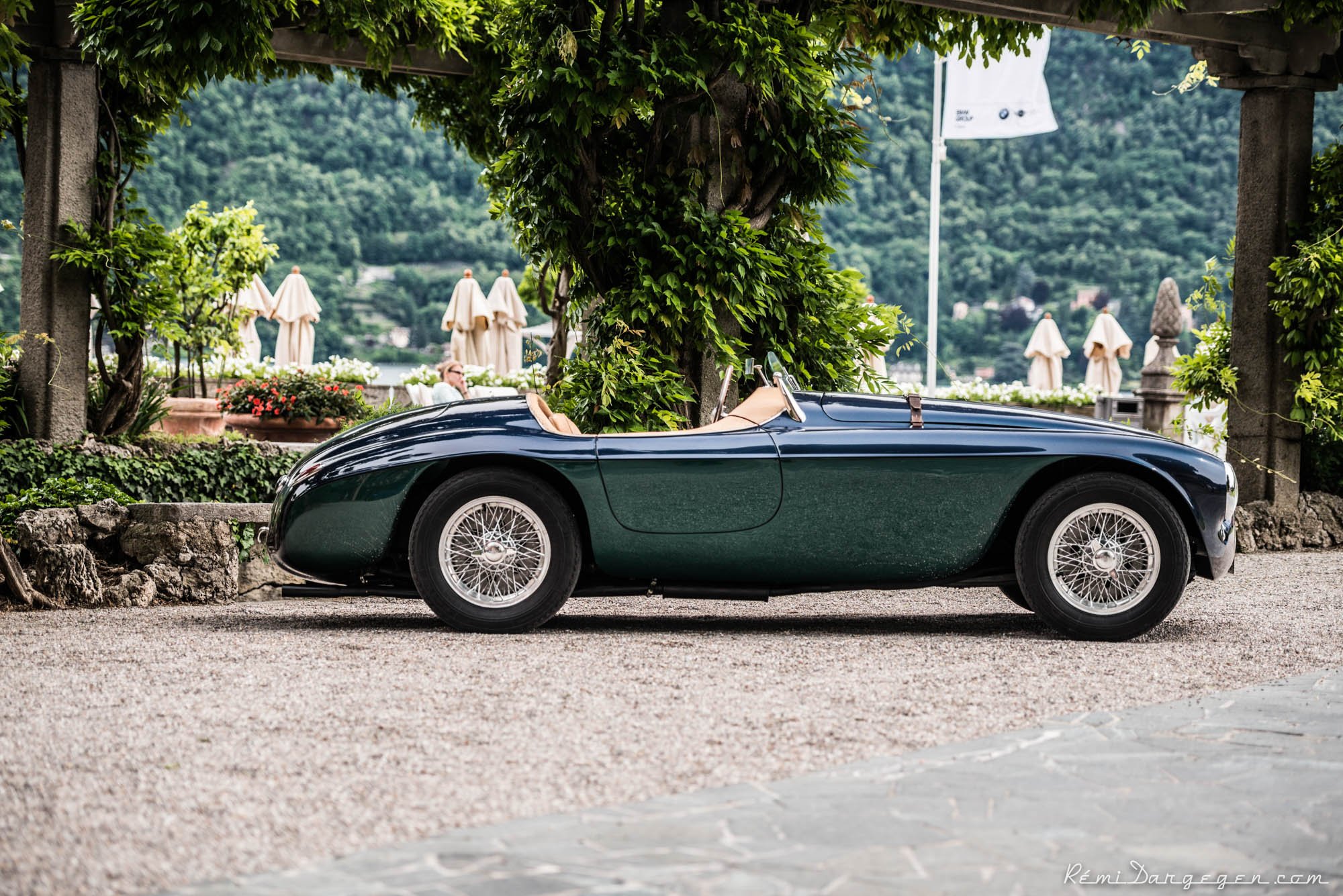 A vintage green convertible sports car parked on a gravel path under a leafy archway with a scenic background of mountains, greenery, and closed umbrellas to the lake.