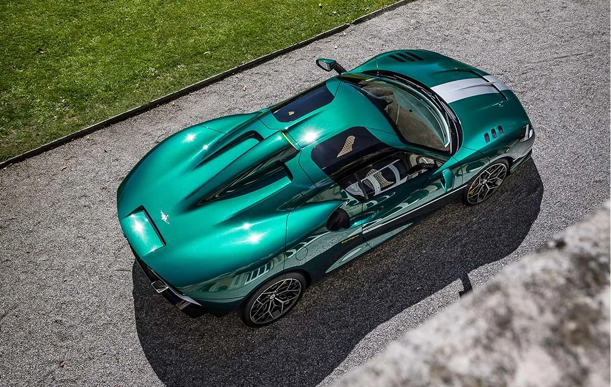 A sleek green sports car with black accents and racing stripes viewed from above on a gravel driveway next to grass.