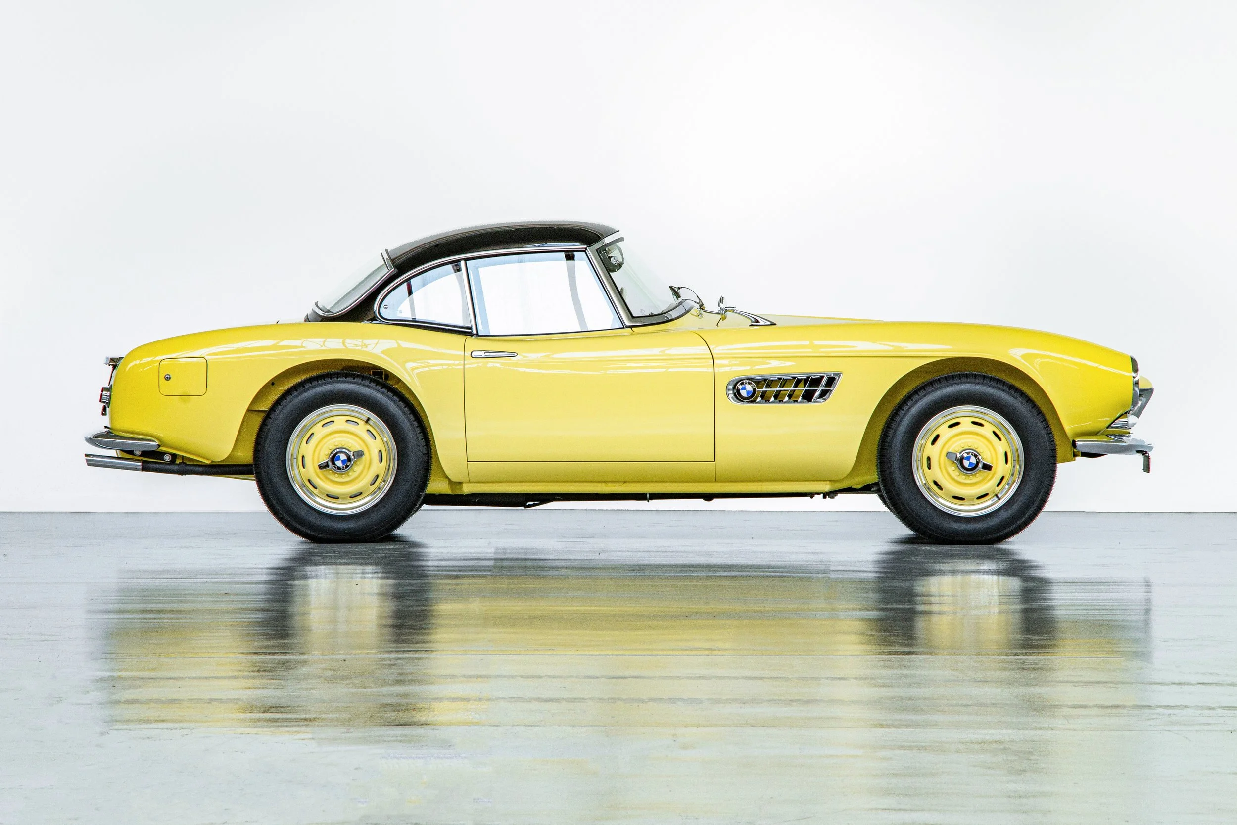 A yellow vintage BMW convertible car with black roof parked on a polished concrete floor against a white wall.