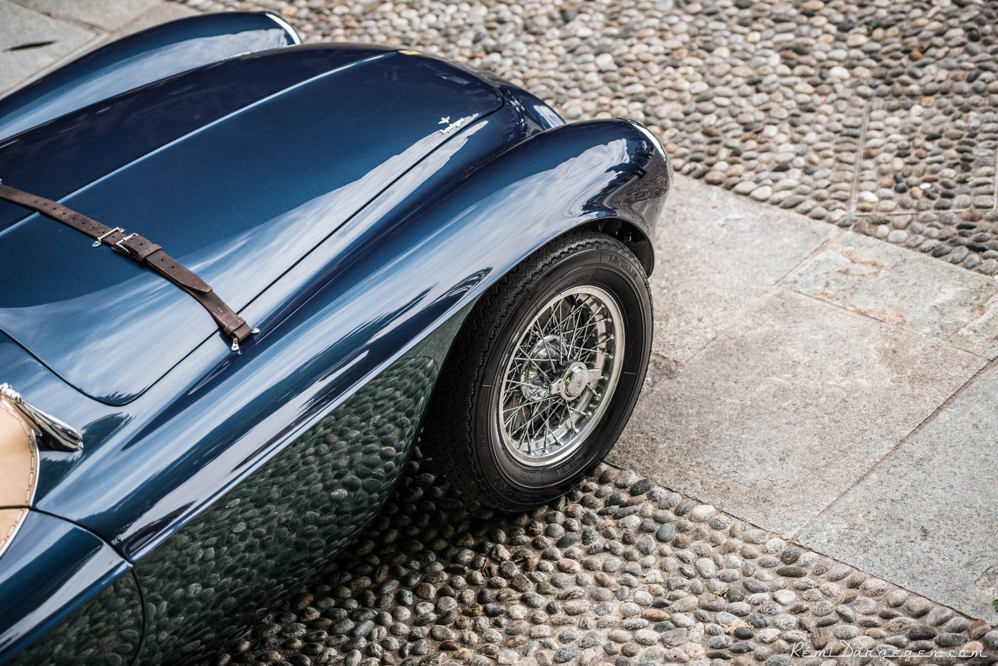 Close-up of a classic dark blue car with wire-spoke wheels, parked on a cobblestone and concrete surface.