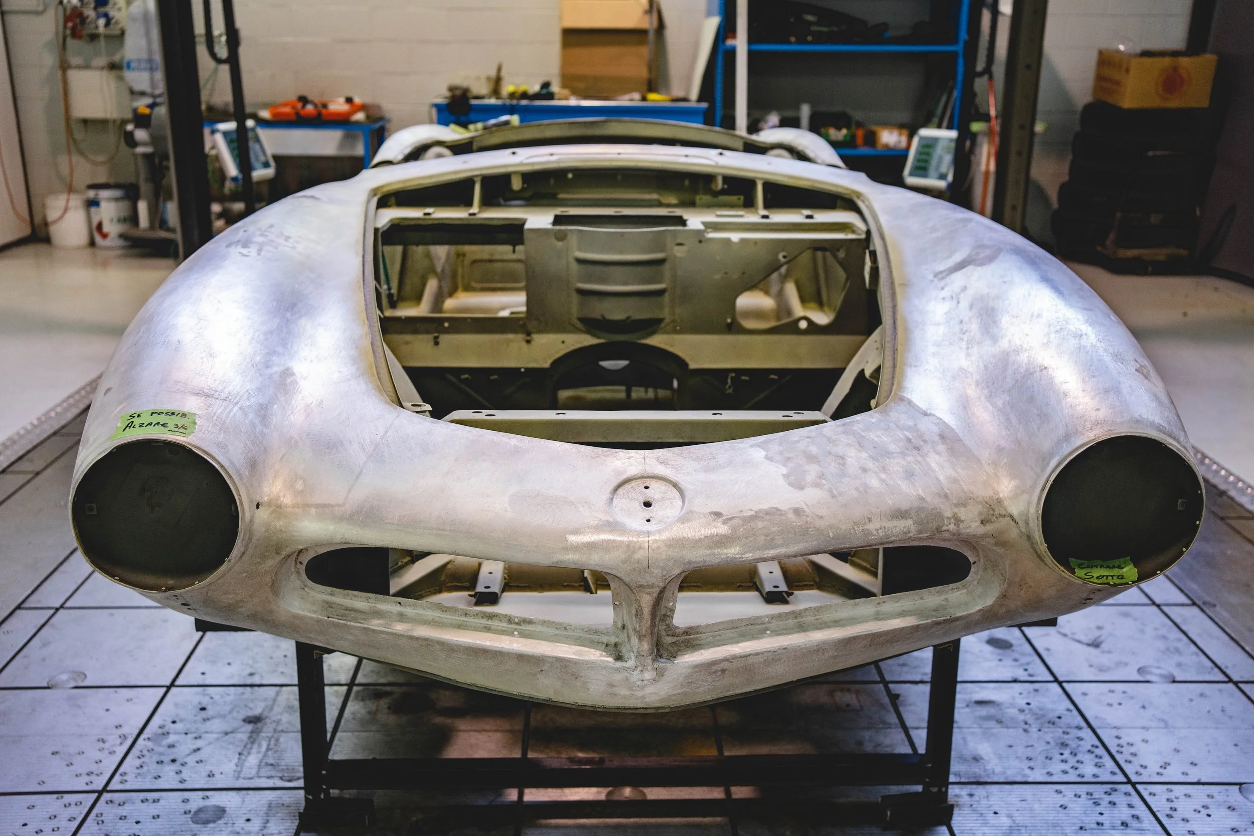 Unfinished vintage race car body shell in a workshop, made of metal, with round openings for headlights and a central opening for the windshield, placed on a stand.
