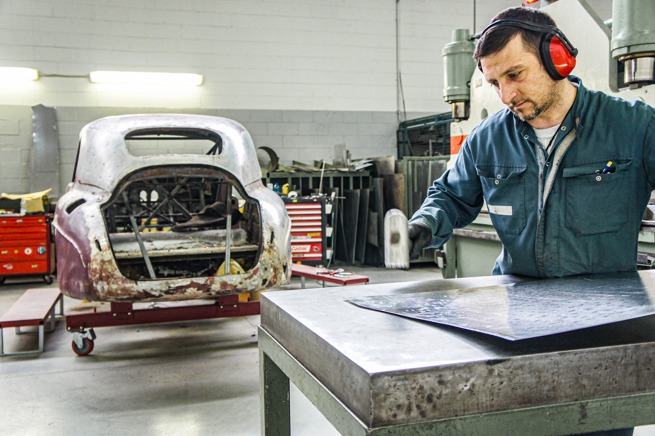 A man wearing ear protection and gloves working on a metal table in an auto repair shop with a rusty, stripped vintage car body in the background.