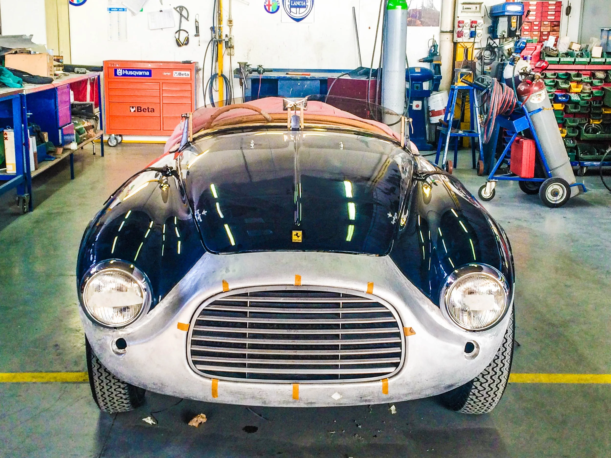Front view of a vintage Ferrari race car inside a workshop with tools and equipment in the background.