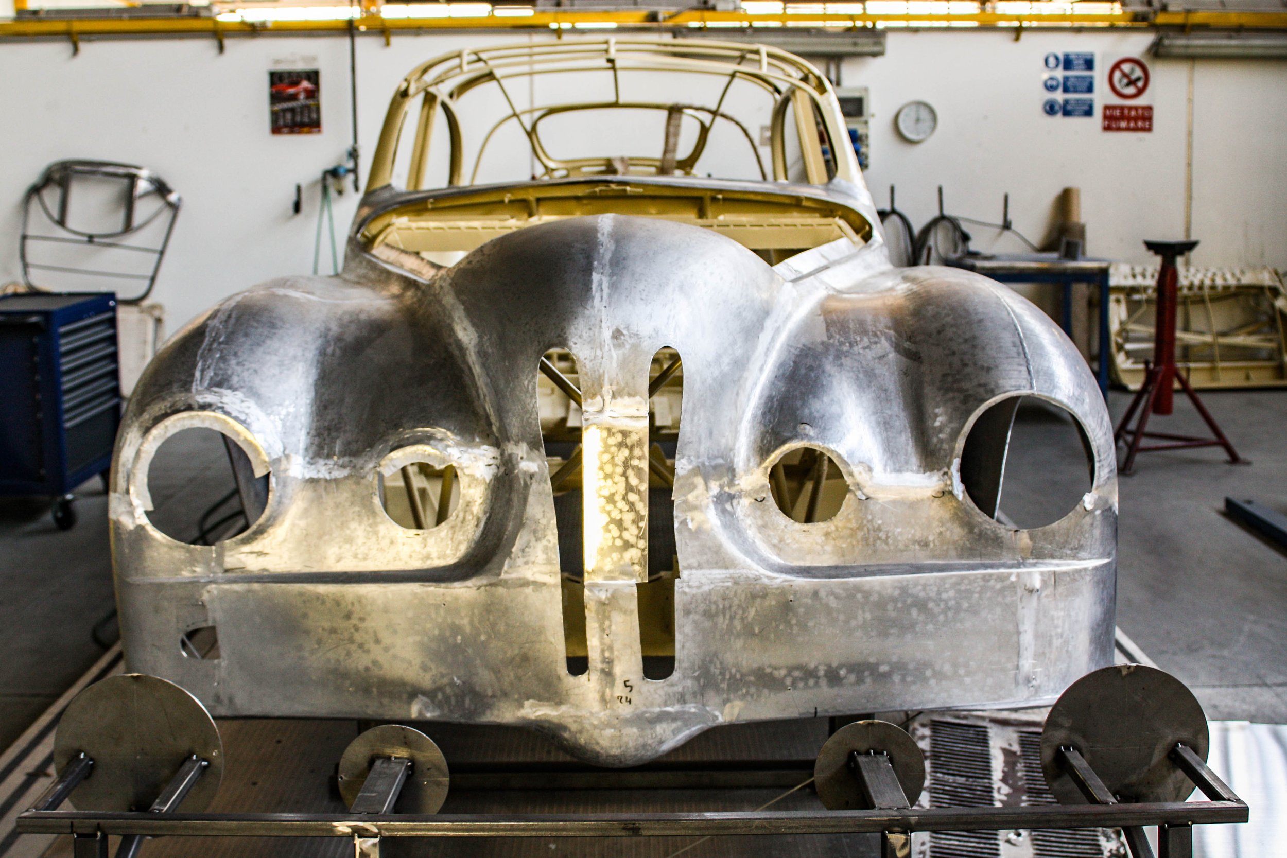 A partially assembled vintage race car body in a workshop, with metal framework and body panels on a wheeled dolly.