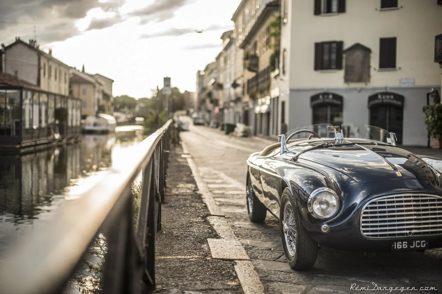 A vintage black sports car with a convertible top parked on a cobblestone street beside a canal, with buildings lining the street and a cloudy sky overhead.