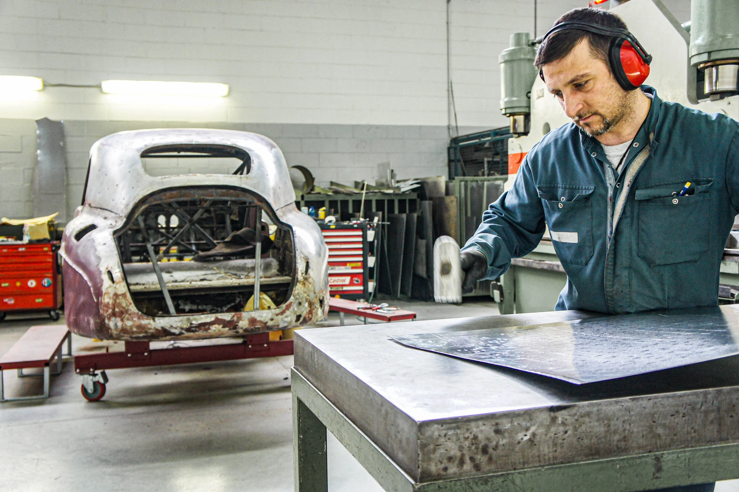 A man working in a metal workshop with a partially restored vintage car body on a cart behind him.