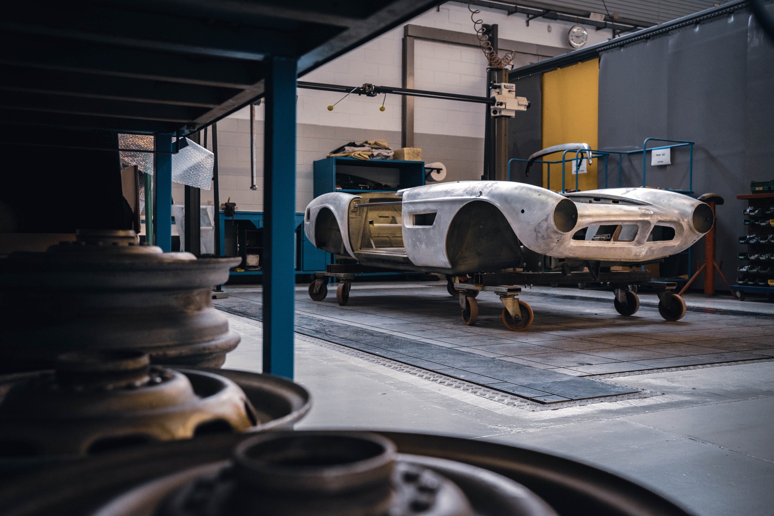 An unfinished vintage race car body on a wheeled cart in an auto workshop, with tooling and equipment around.