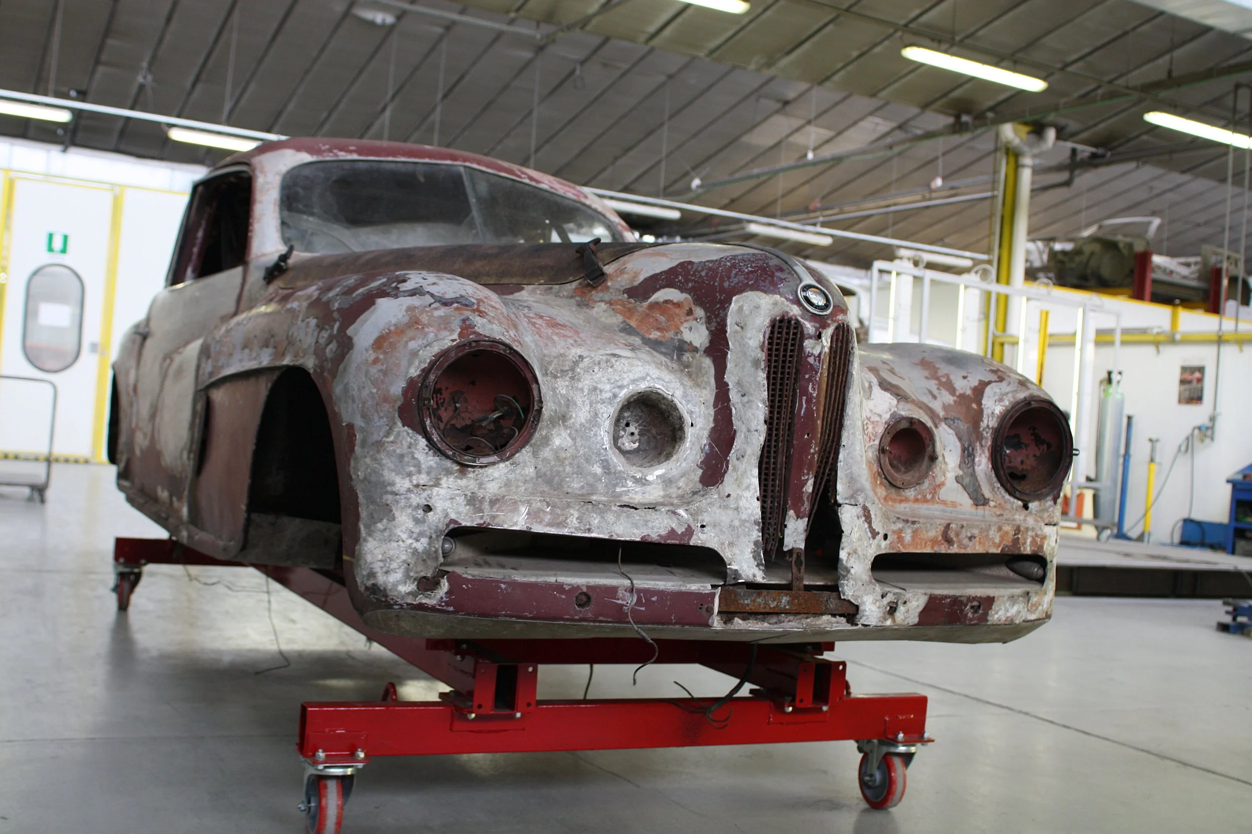 A vintage car shell undergoing restoration in a workshop, with visible rust and peeling paint, mounted on a red rolling platform.