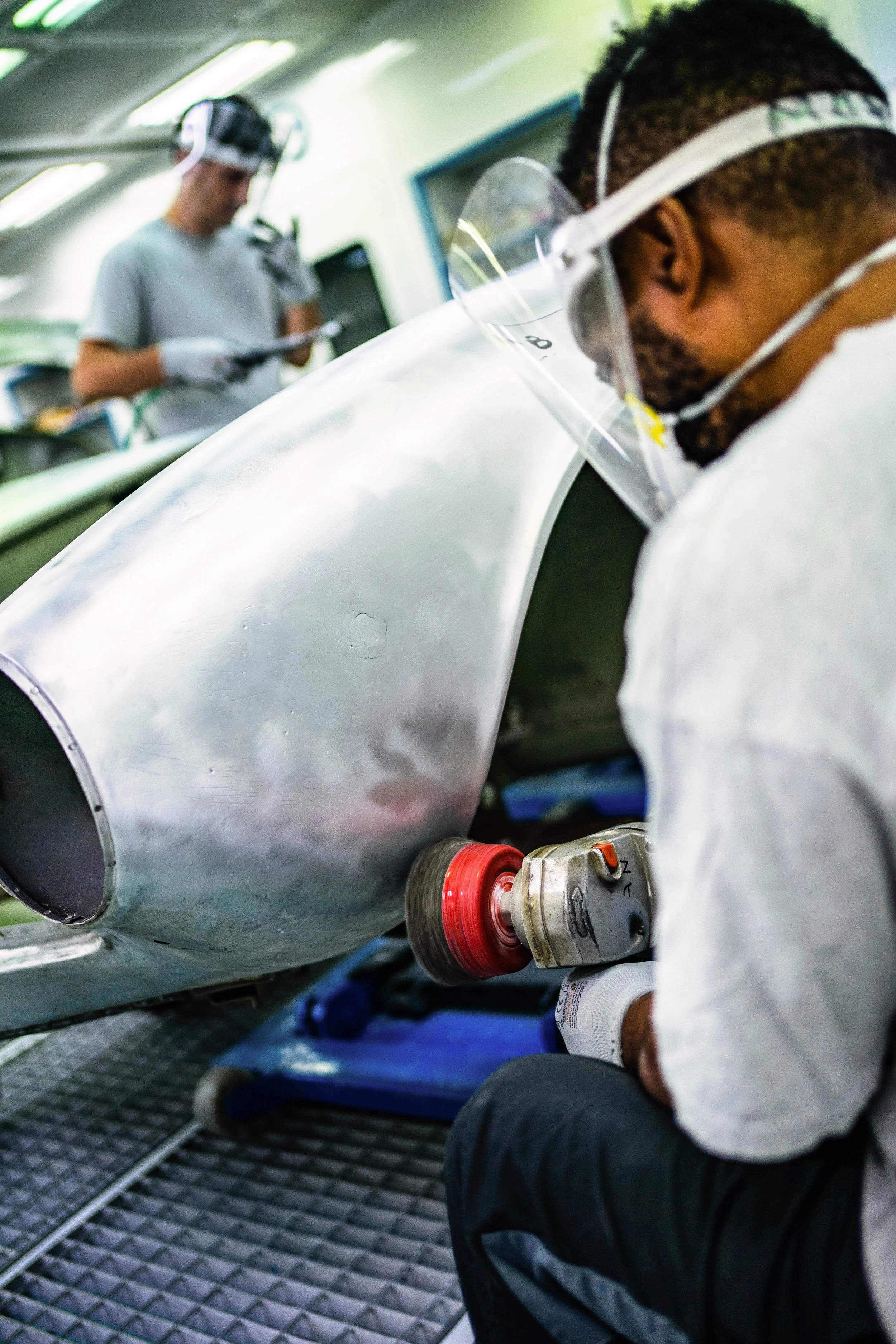 A man wearing safety glasses and gloves polishes a large metal aircraft part with a handheld polishing tool, while another person working in the background inside a workshop.