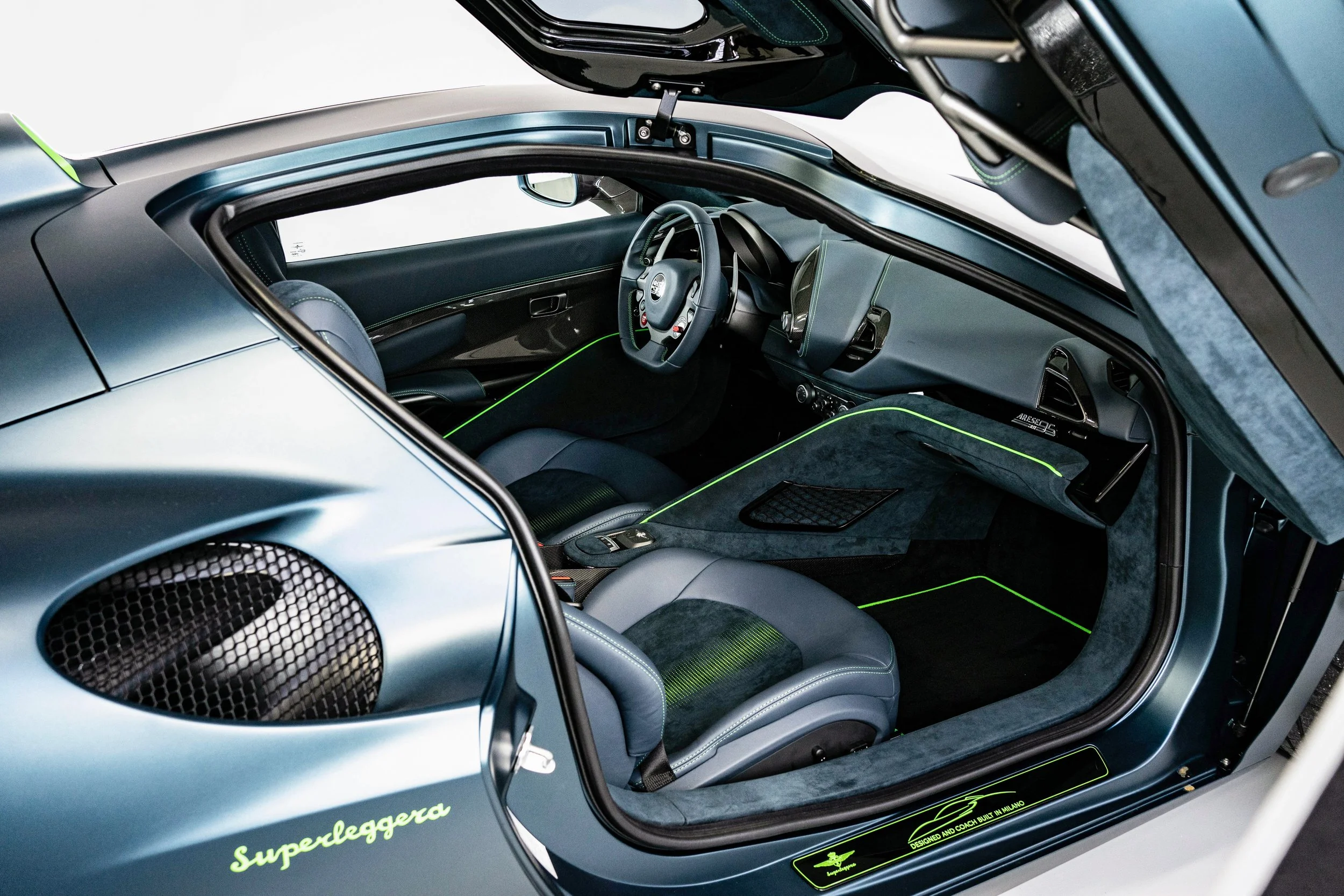 Interior of a luxury sports car showing racing seats, steering wheel, dashboard, and open gull-wing door with branding and neon green accents.